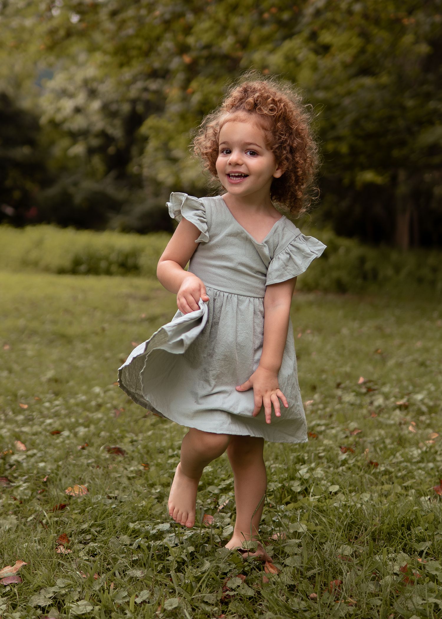 Curly-haired toddler girl in sage green dress twirling barefoot in green grass.