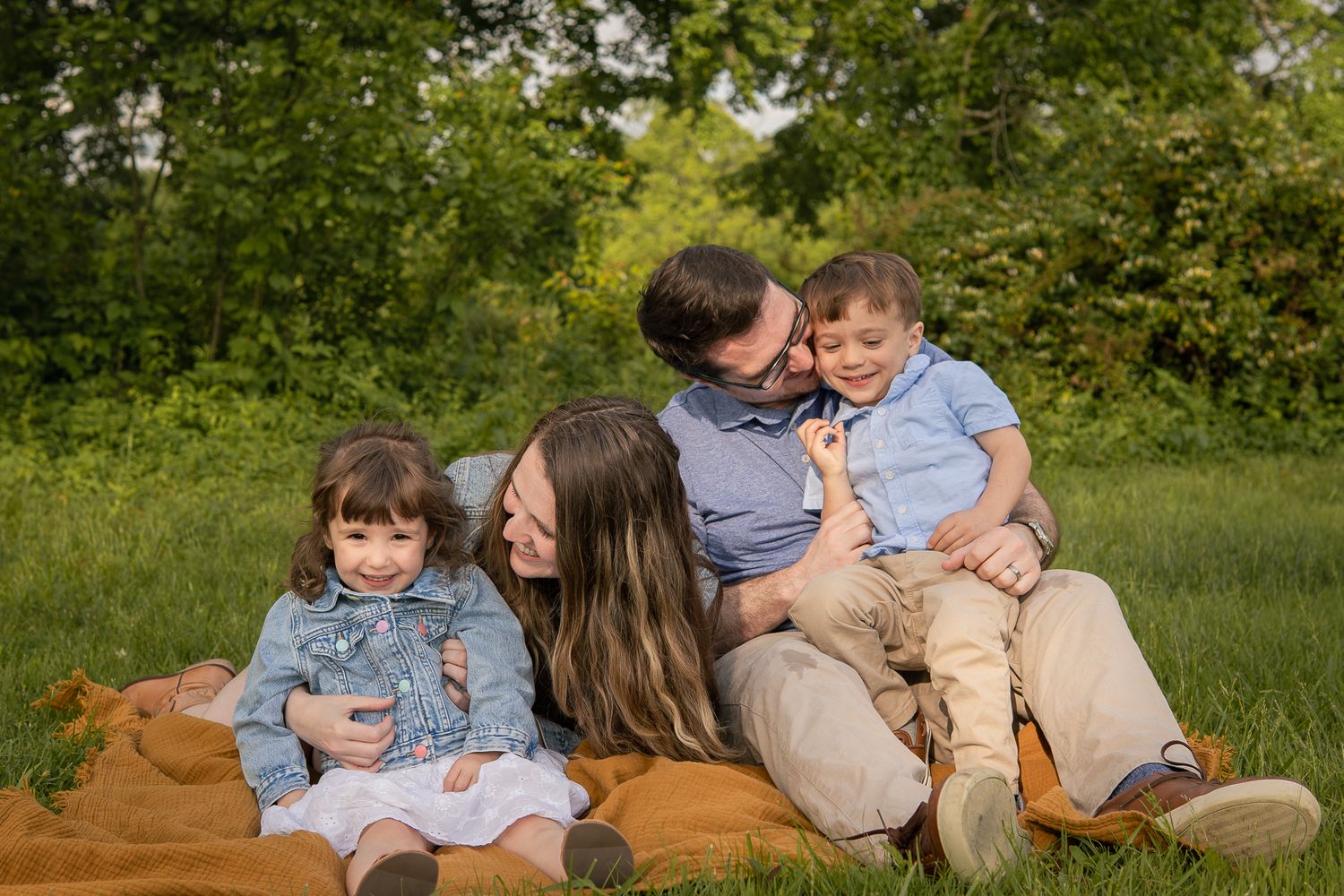 Family sitting together on a brown blanket outdoors surrounded by green grass and trees.