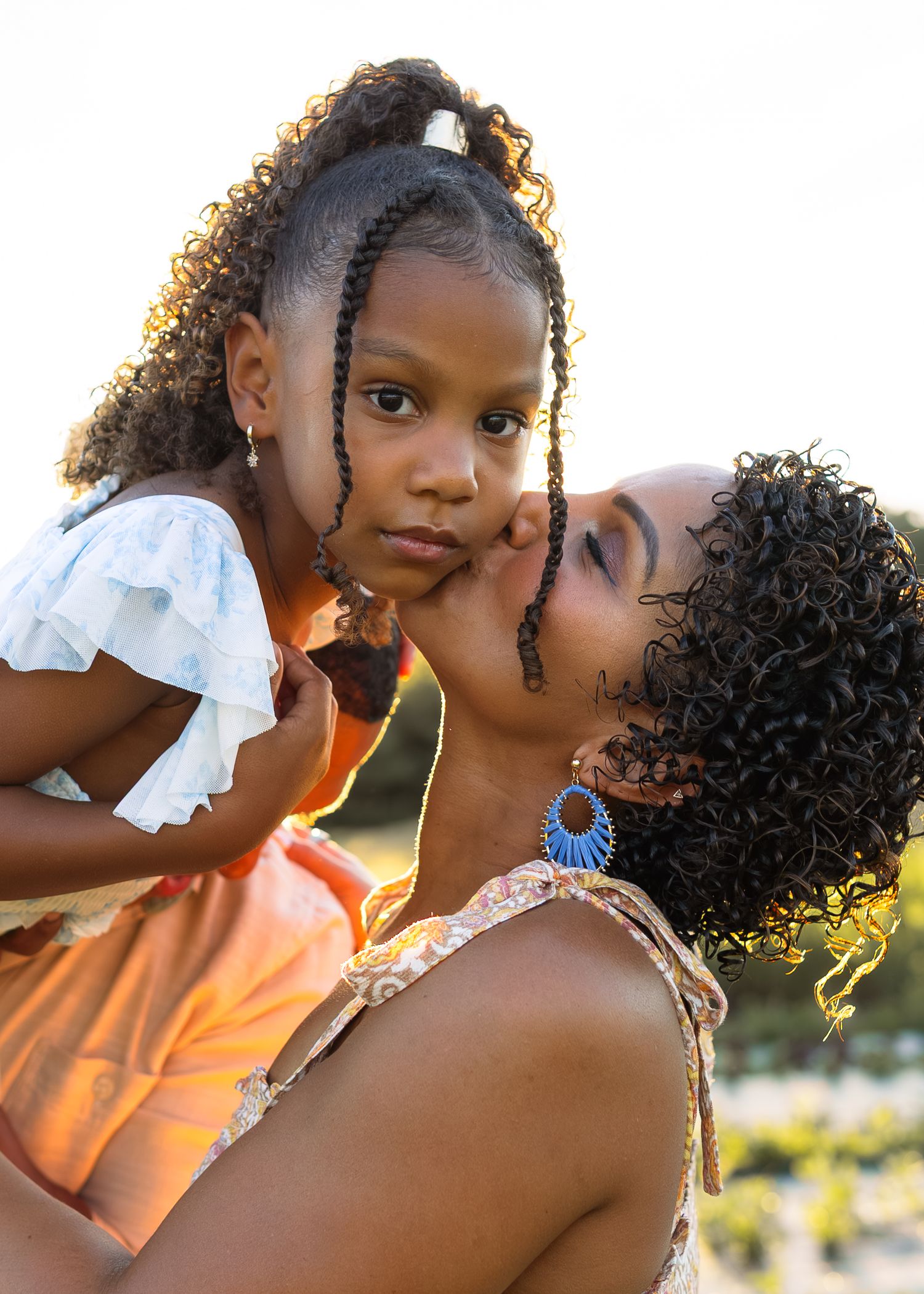 A mother tenderly kisses her young daughter's cheek while holding her up outdoors in warm golden light.