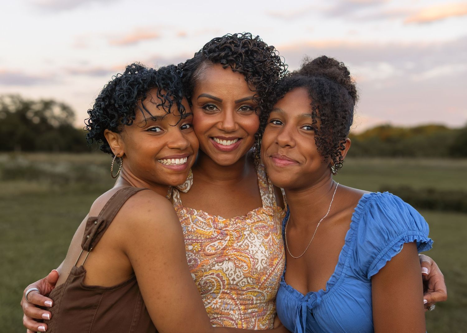 Three smiling women with curly hair pose together outdoors at sunset in a green field, wearing colorful summer outfits.