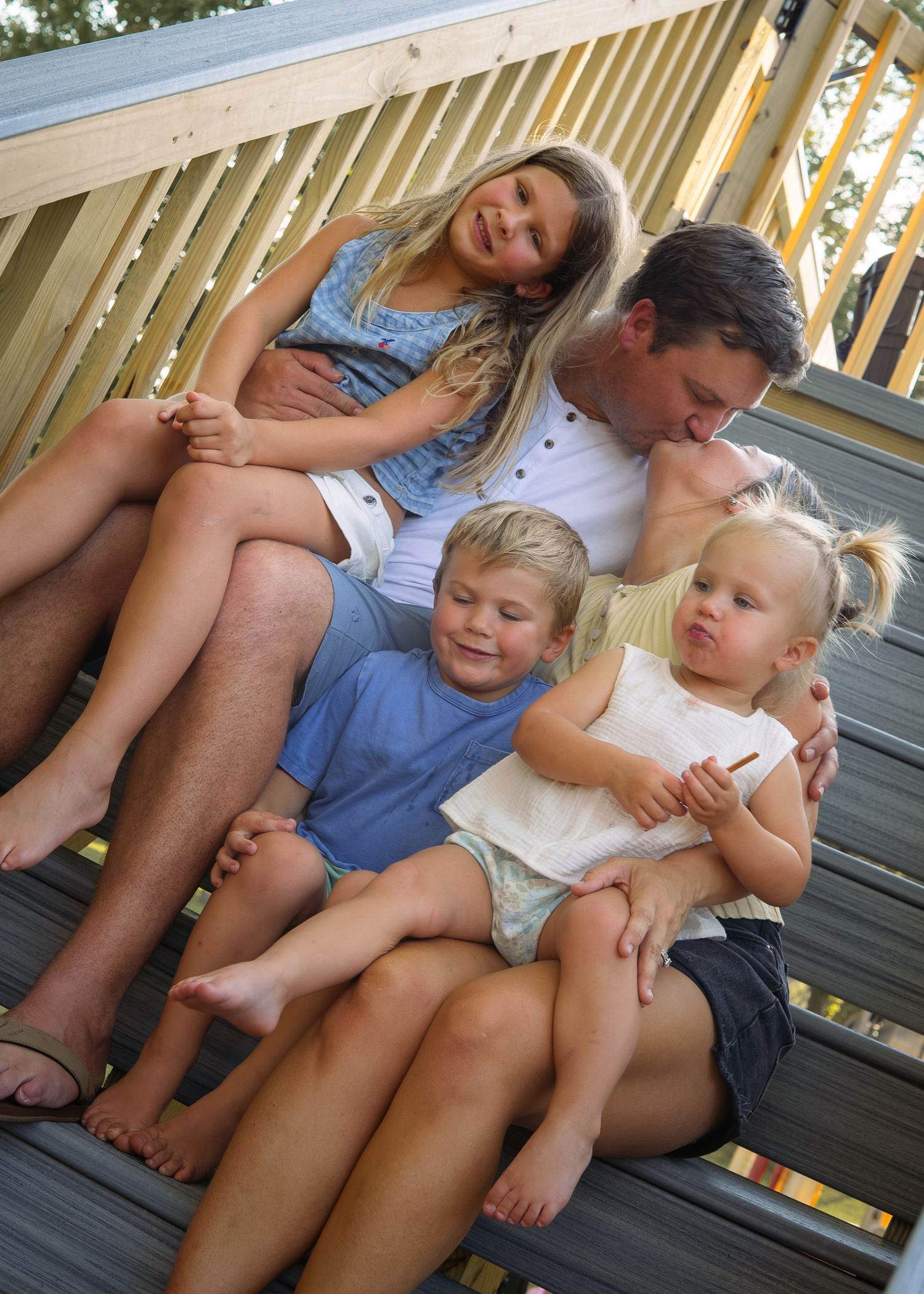 Happy family of five sitting together on wooden deck steps, parents embracing three young children outdoors.