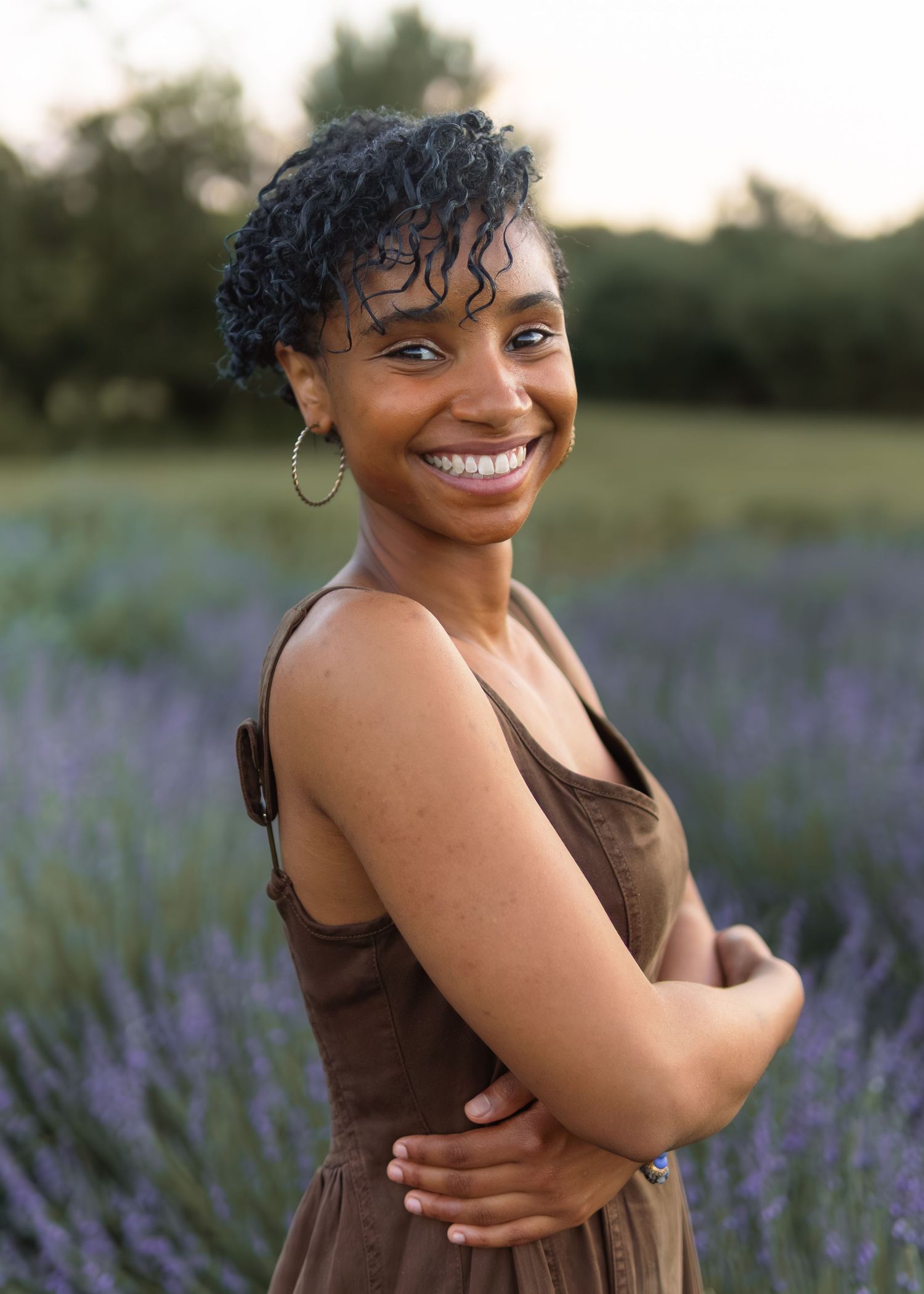 Smiling young woman in brown dress with arms crossed in lavender field at sunset.