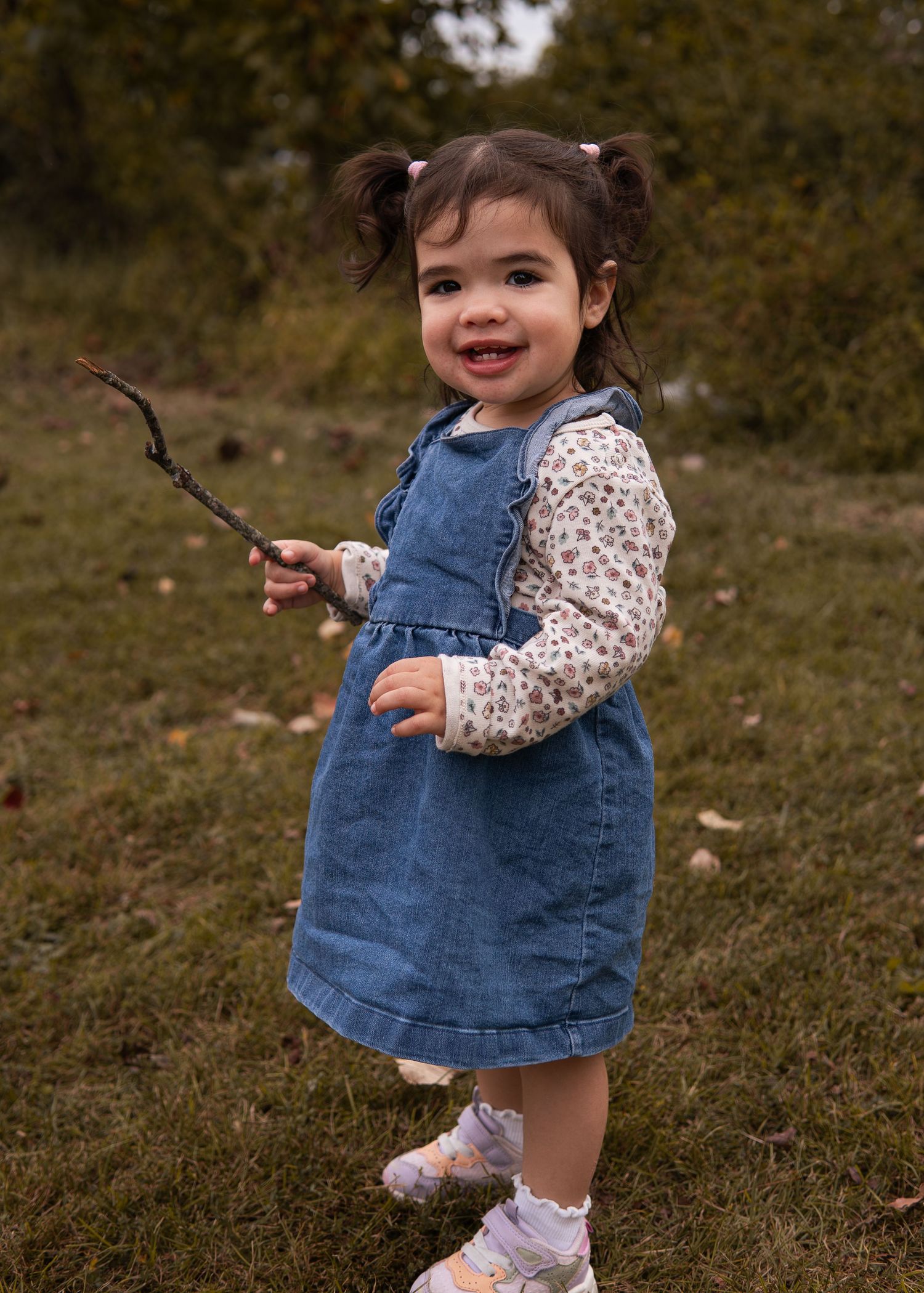 Toddler girl in denim pinafore dress holding a stick outdoors in autumn.