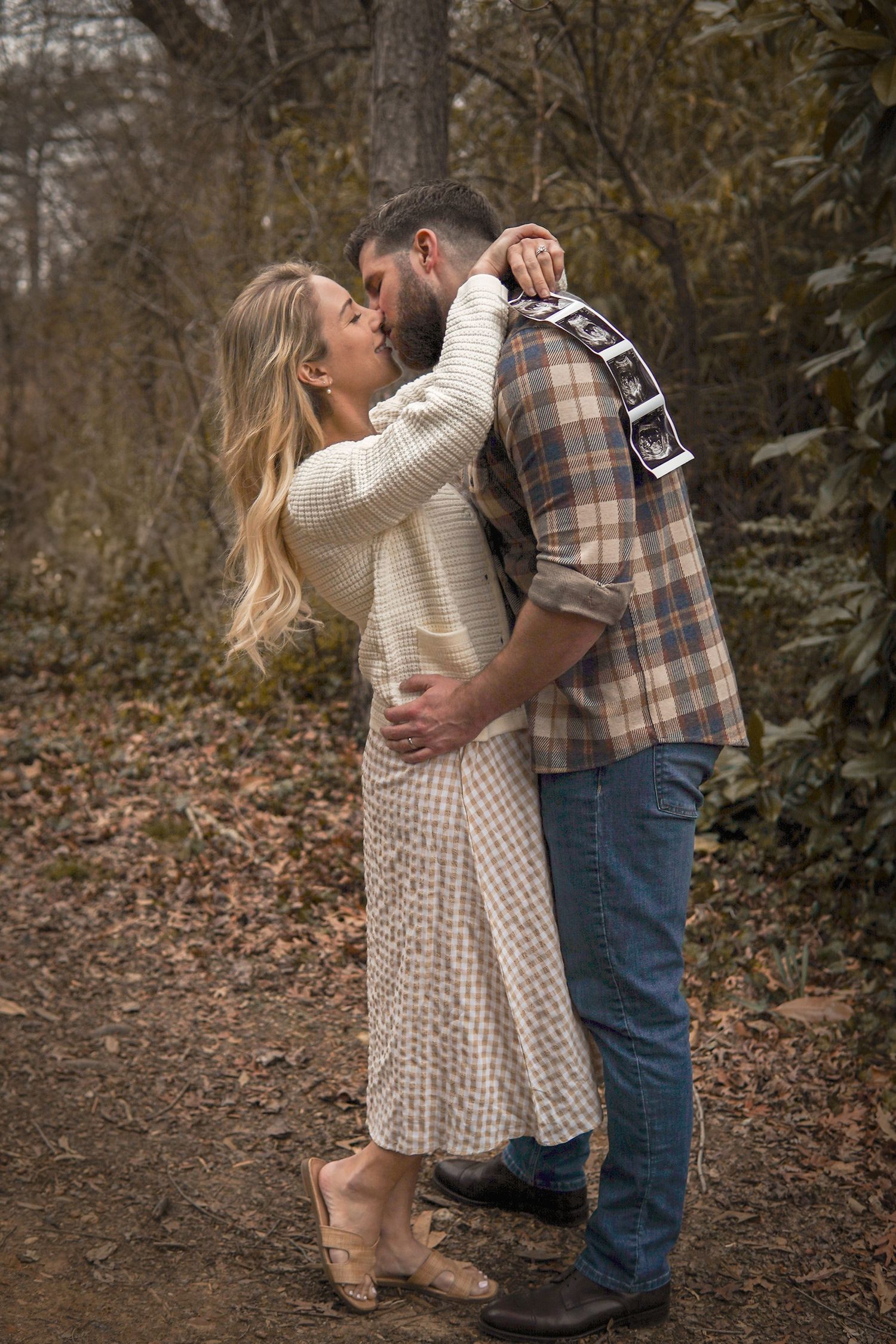 A couple embraces on a wooded path in autumn, wearing cozy sweaters and sharing an intimate moment together.