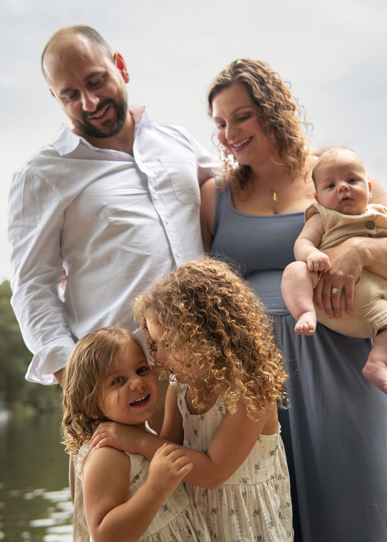 A happy family of five poses outdoors near water, with parents holding a newborn and two young curly-haired girls hugging.