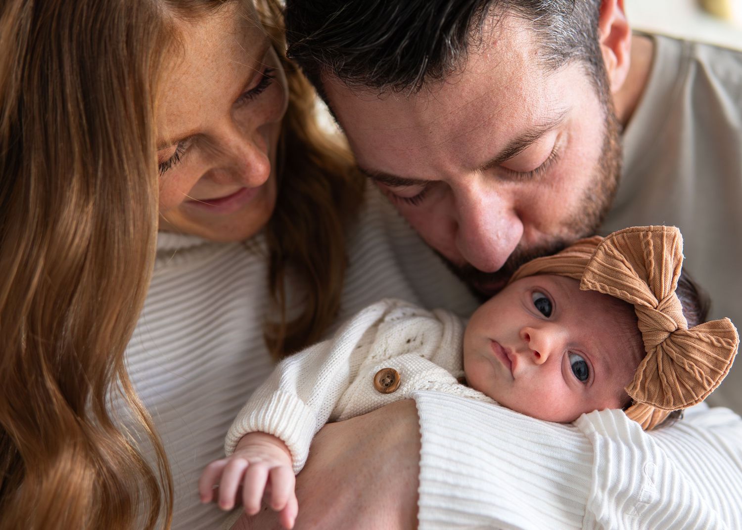 New parents lovingly gazing at their newborn baby wrapped in a white blanket with a bow headband.