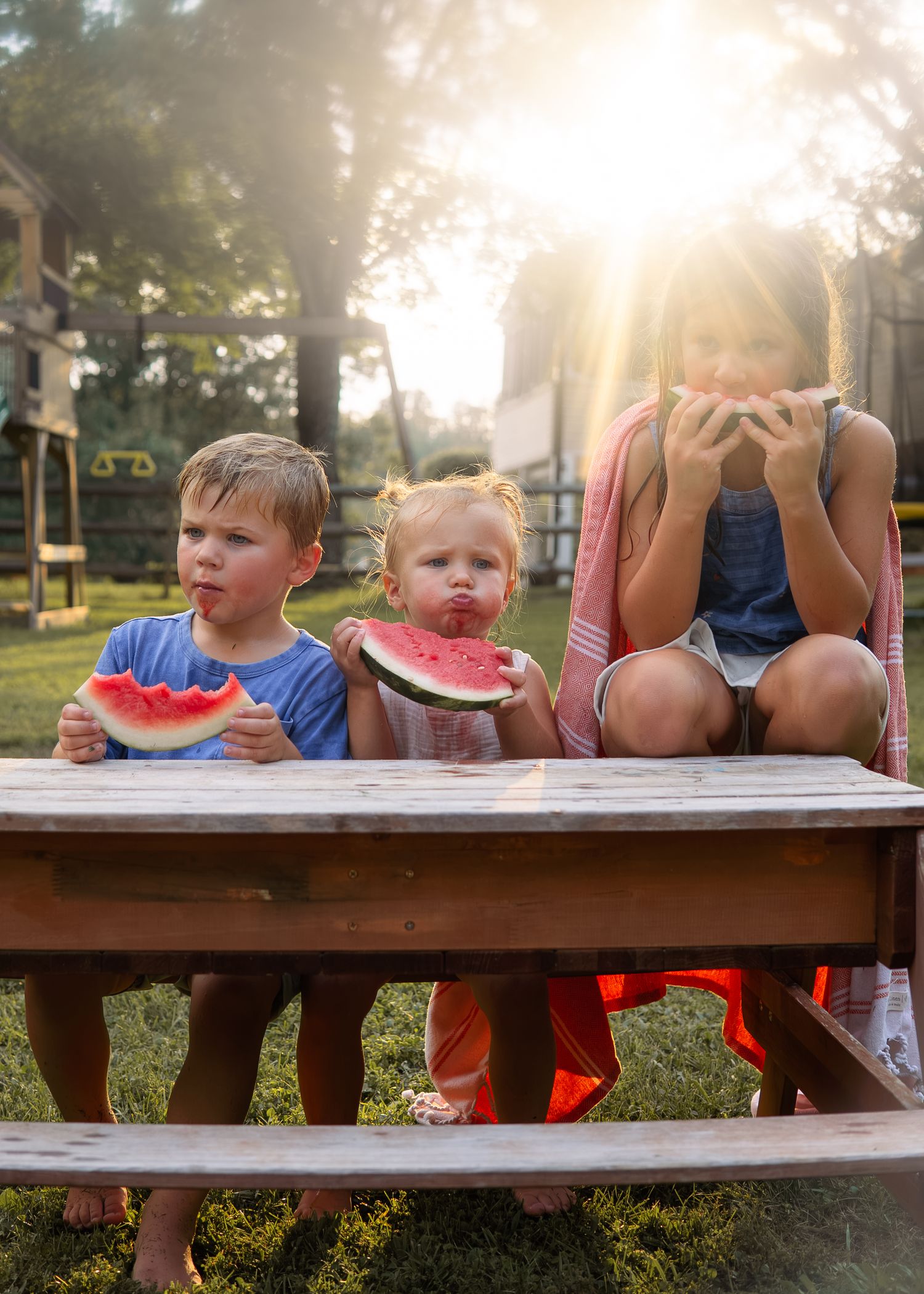 Three children eating watermelon slices at a wooden picnic table outdoors in golden sunlight.