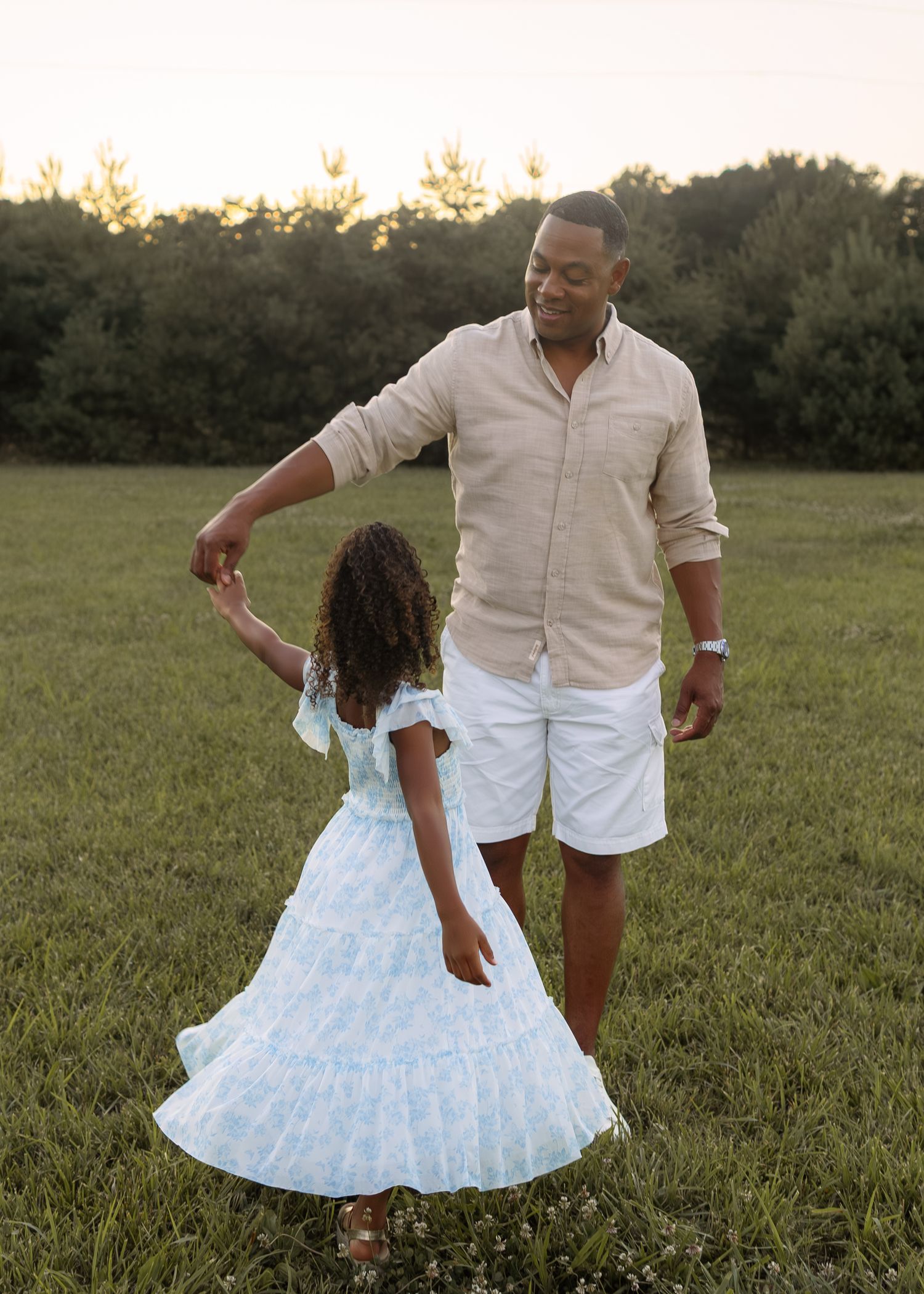 A father dances with his young daughter in a green field at sunset, both smiling joyfully.