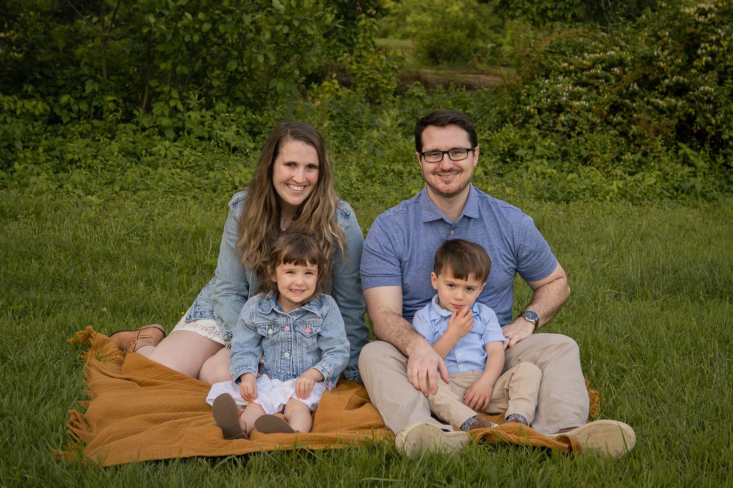 A family of four sits together on a brown blanket in a green grassy field during an outdoor photo session.