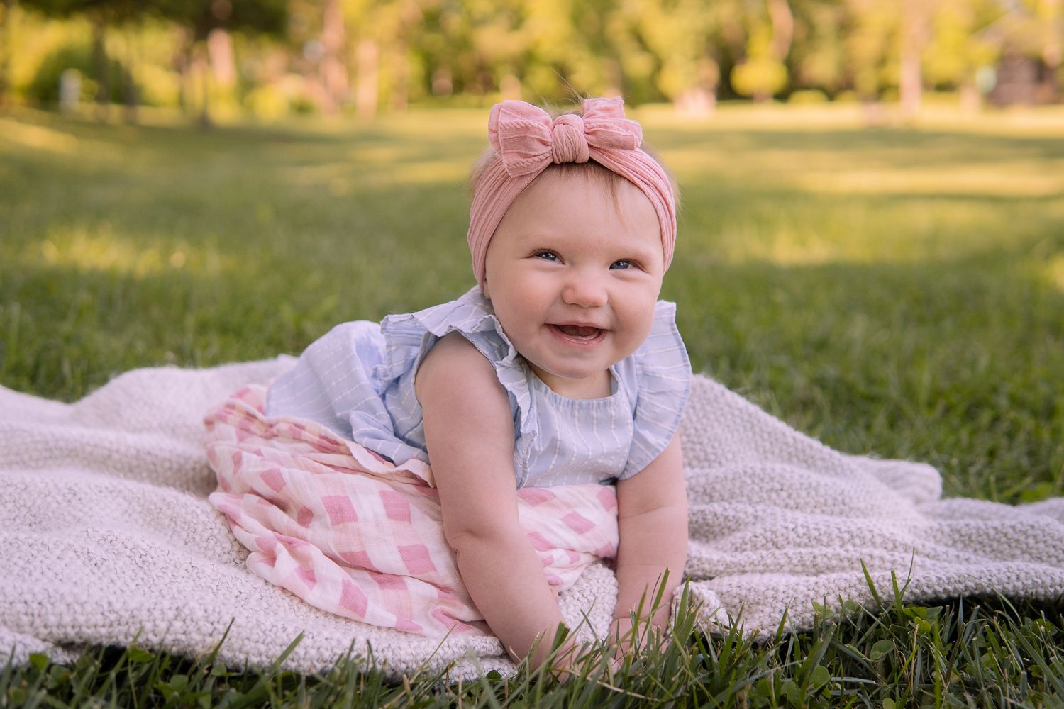 Baby in pink bow headband and pink dress smiles while laying on blanket in sunny park.