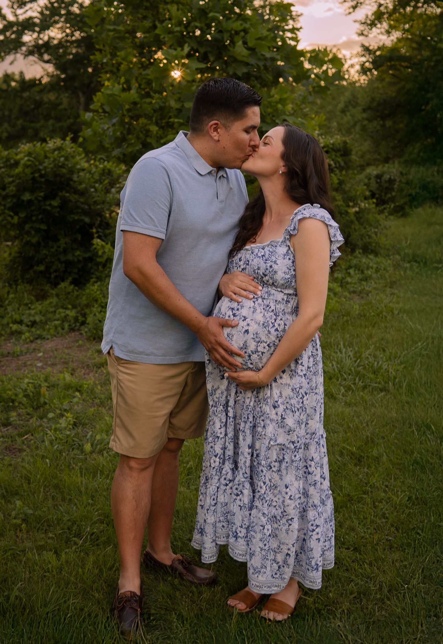 A couple shares a tender moment outdoors at sunset as they display a pregnancy bump in a floral maxi dress and casual attire.