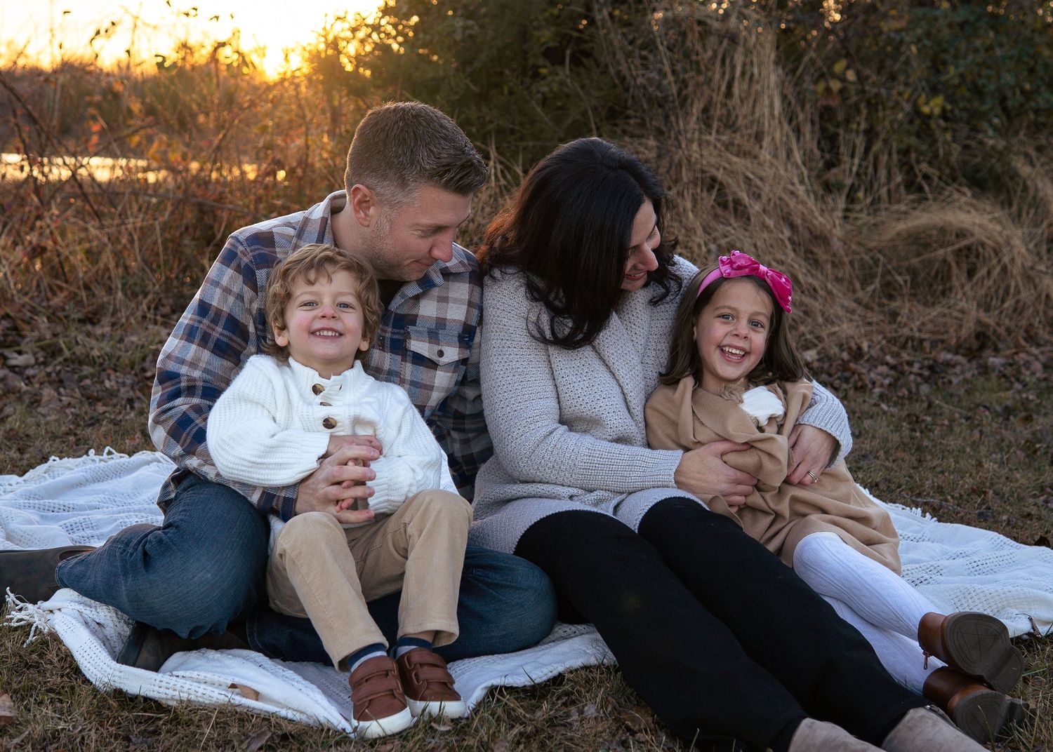 Family of four sitting on blanket laughing together at golden hour outdoors.