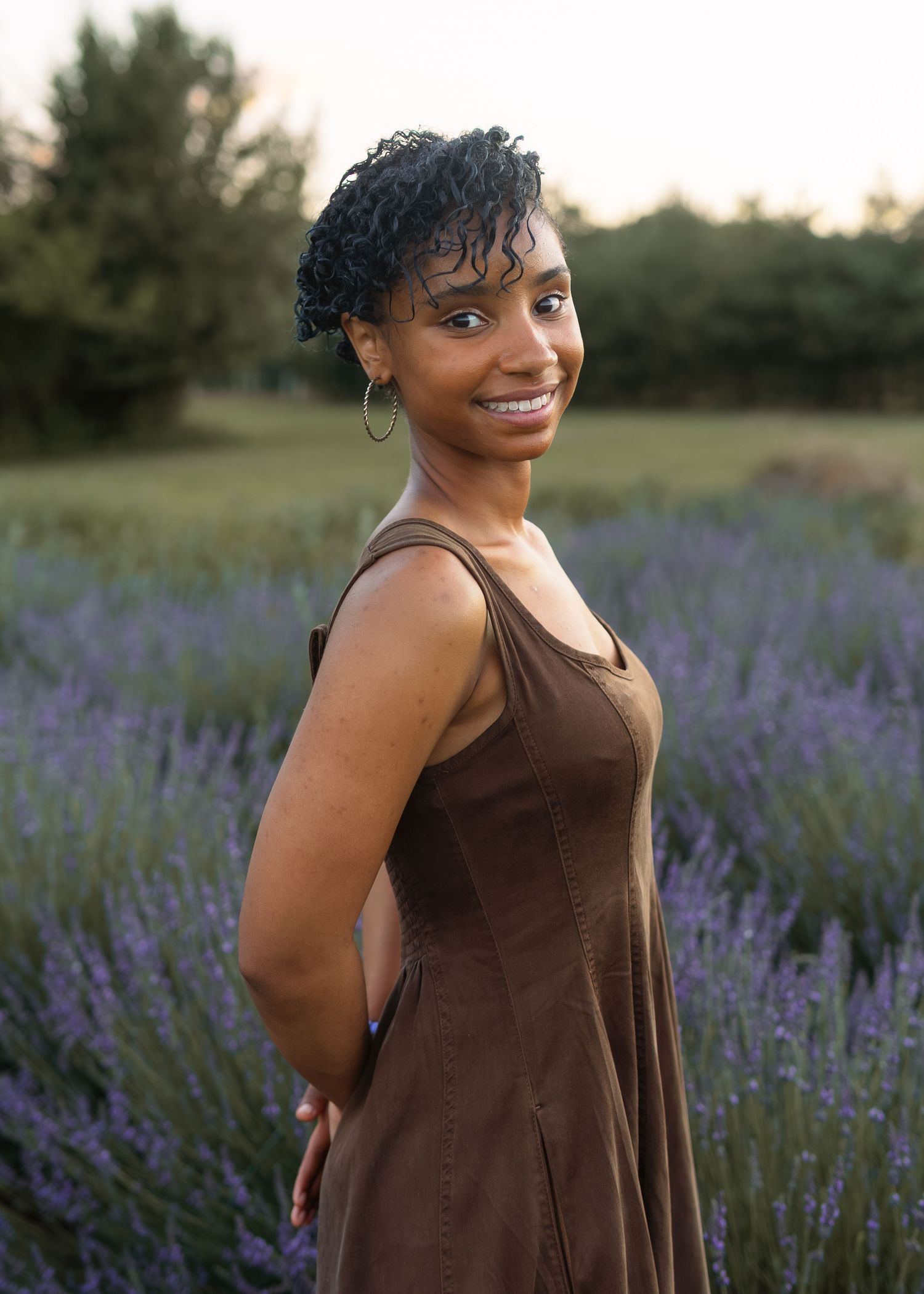 Young woman in brown sundress posing in blooming lavender field during golden hour.