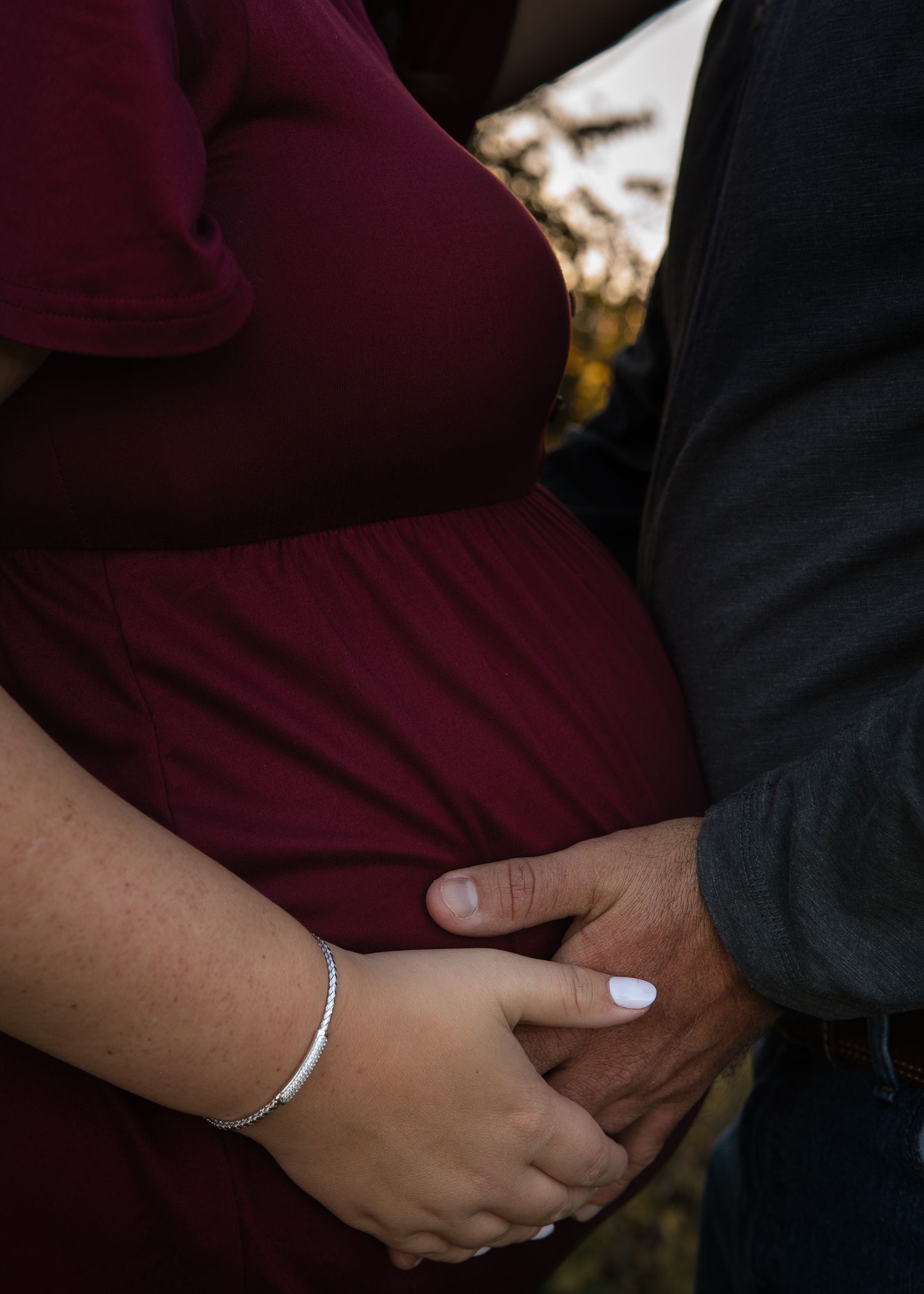 Pregnant woman in burgundy dress holds hands with partner during maternity photoshoot outdoors in autumn.
