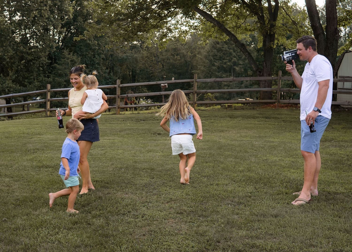 Family playing outdoors on green lawn, man filming with camera as woman holds toddler while two children run toward him.