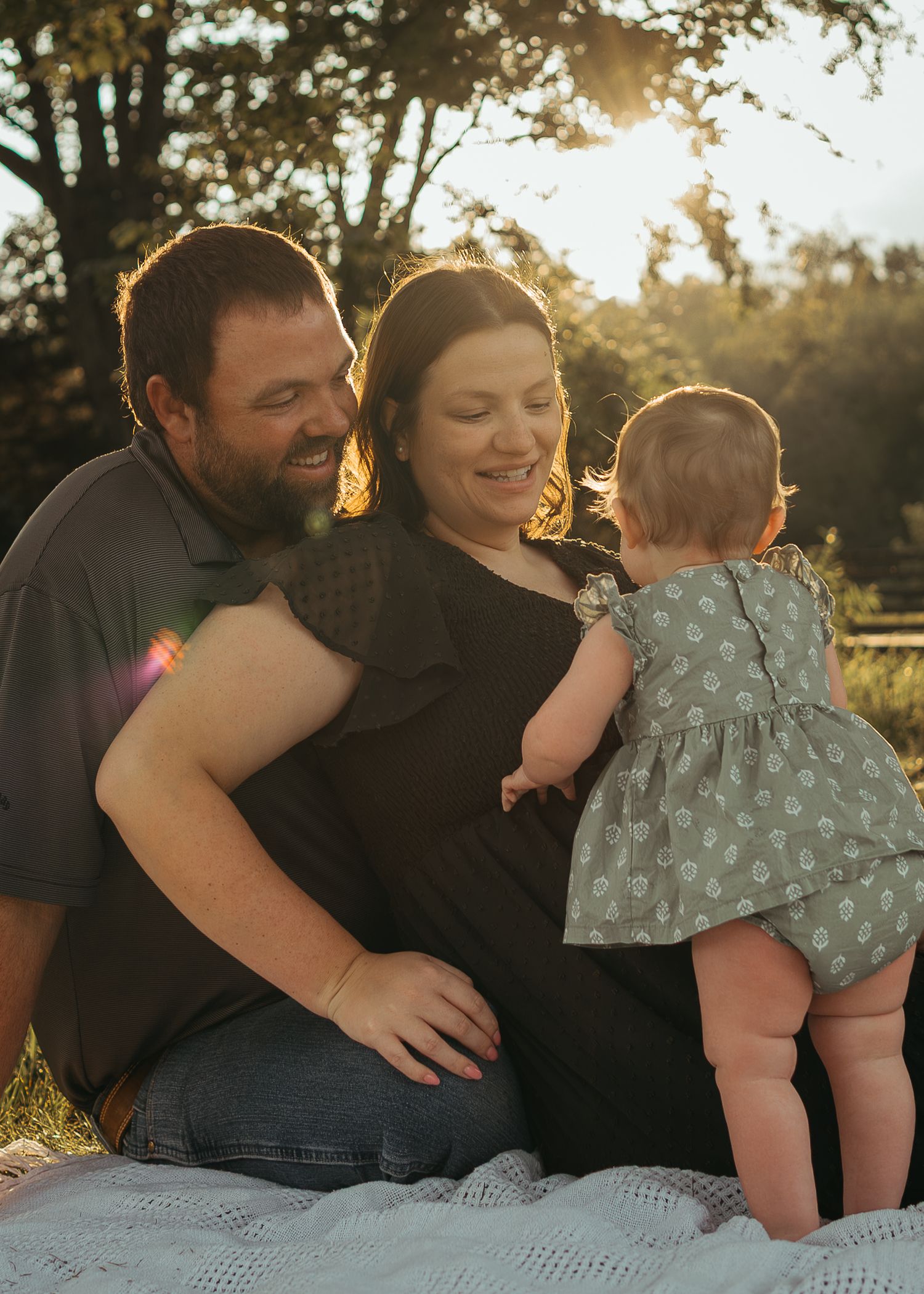 Happy family with baby girl sits together in warm sunset glow on blanket outdoors.