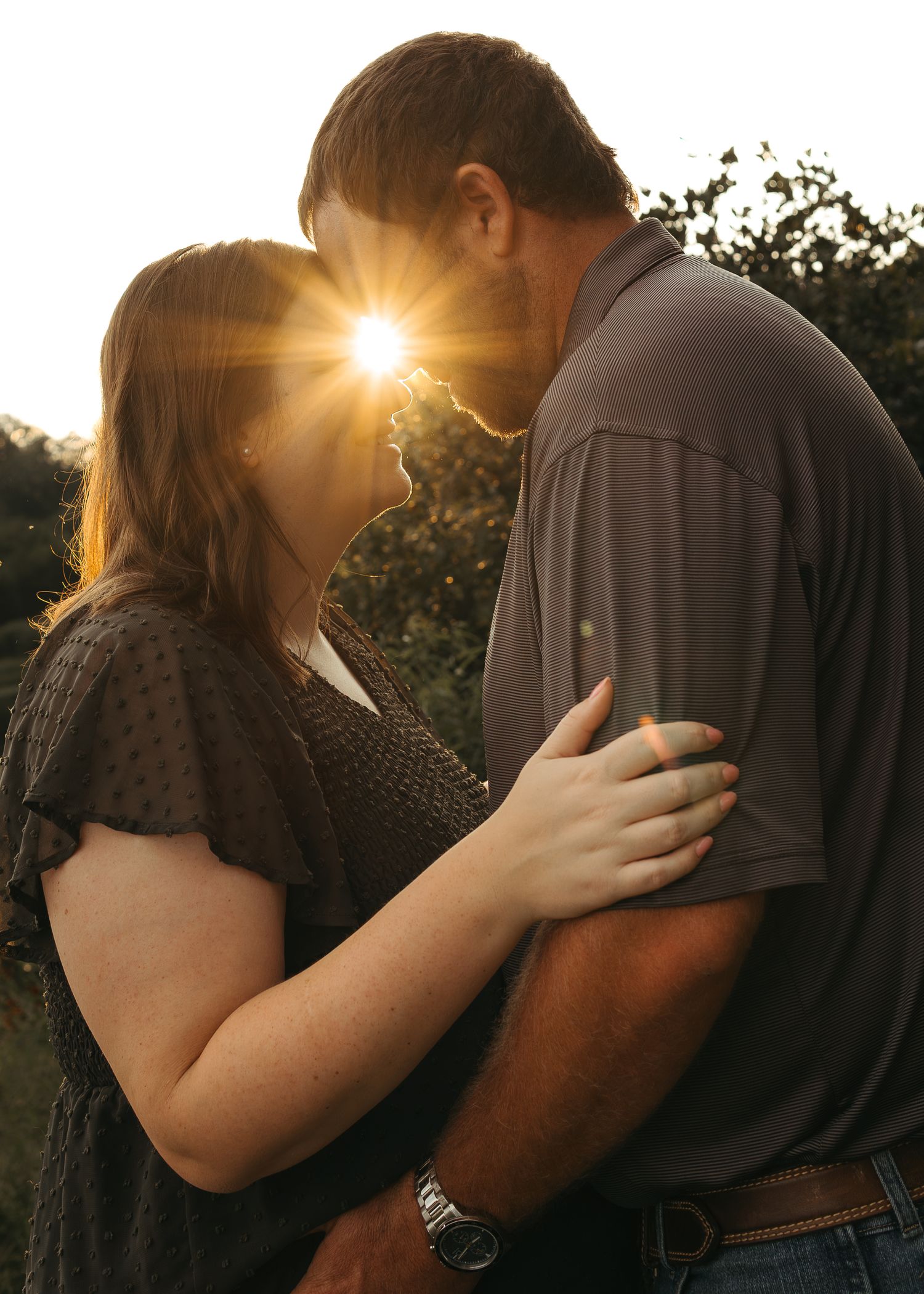 Romantic couple embracing at sunset with golden lens flare between them in an outdoor setting.