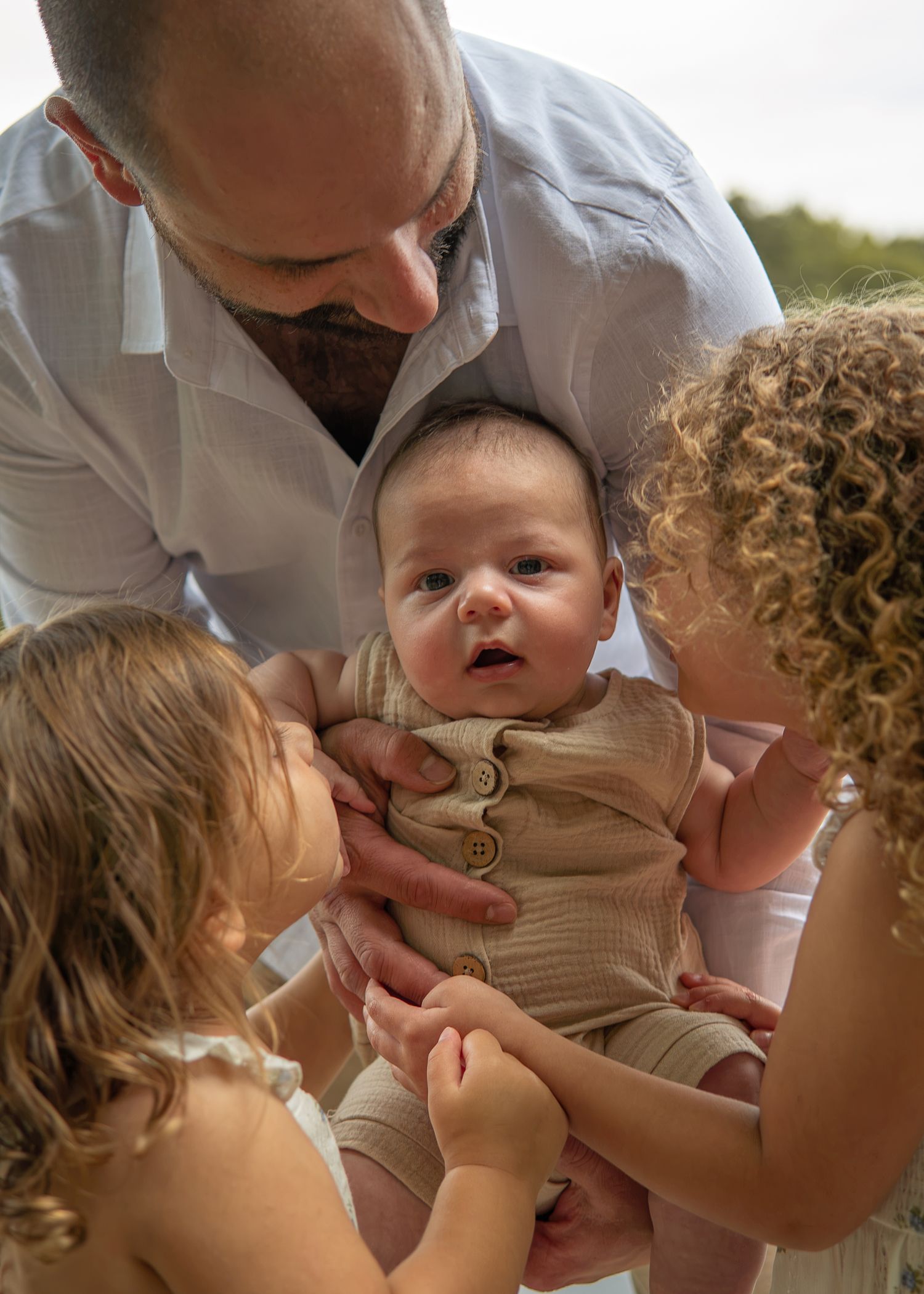 A father holds a newborn baby while two young children lean in close with curiosity and affection outdoors.
