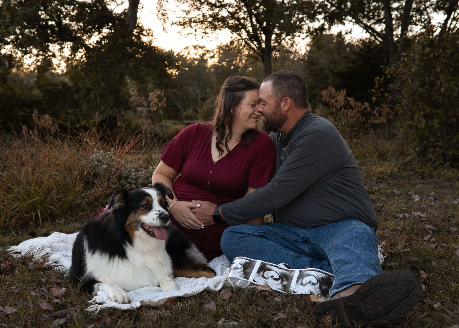 Pregnant couple sitting on blanket with Bernese Mountain Dog at golden hour in autumn field.