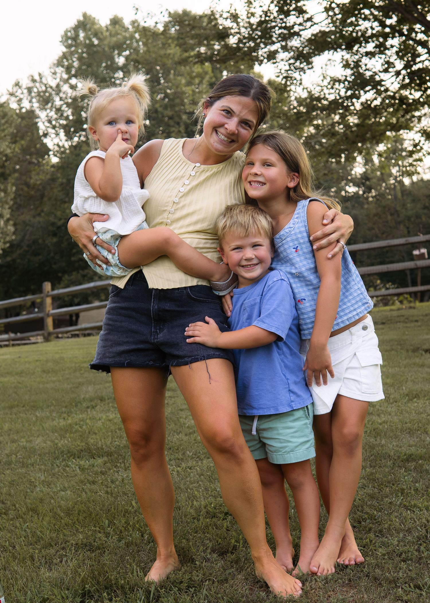 A smiling mother holds a toddler while two children hug her outdoors on green grass near a wooden fence.