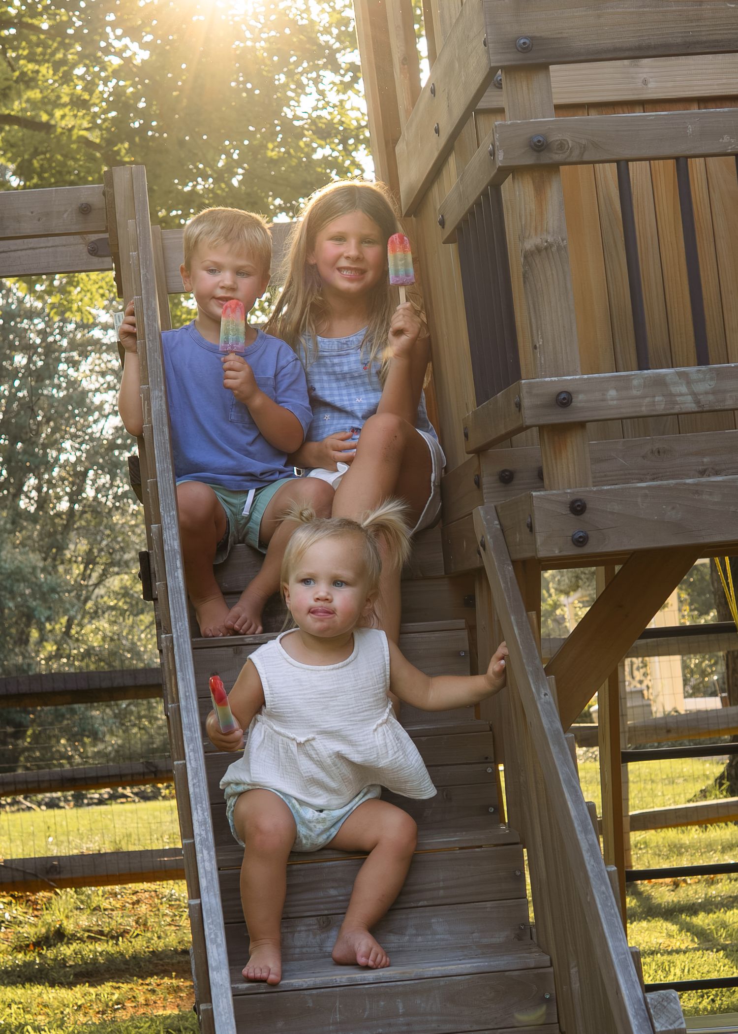 Three children eating popsicles on wooden playground steps in golden sunlight.