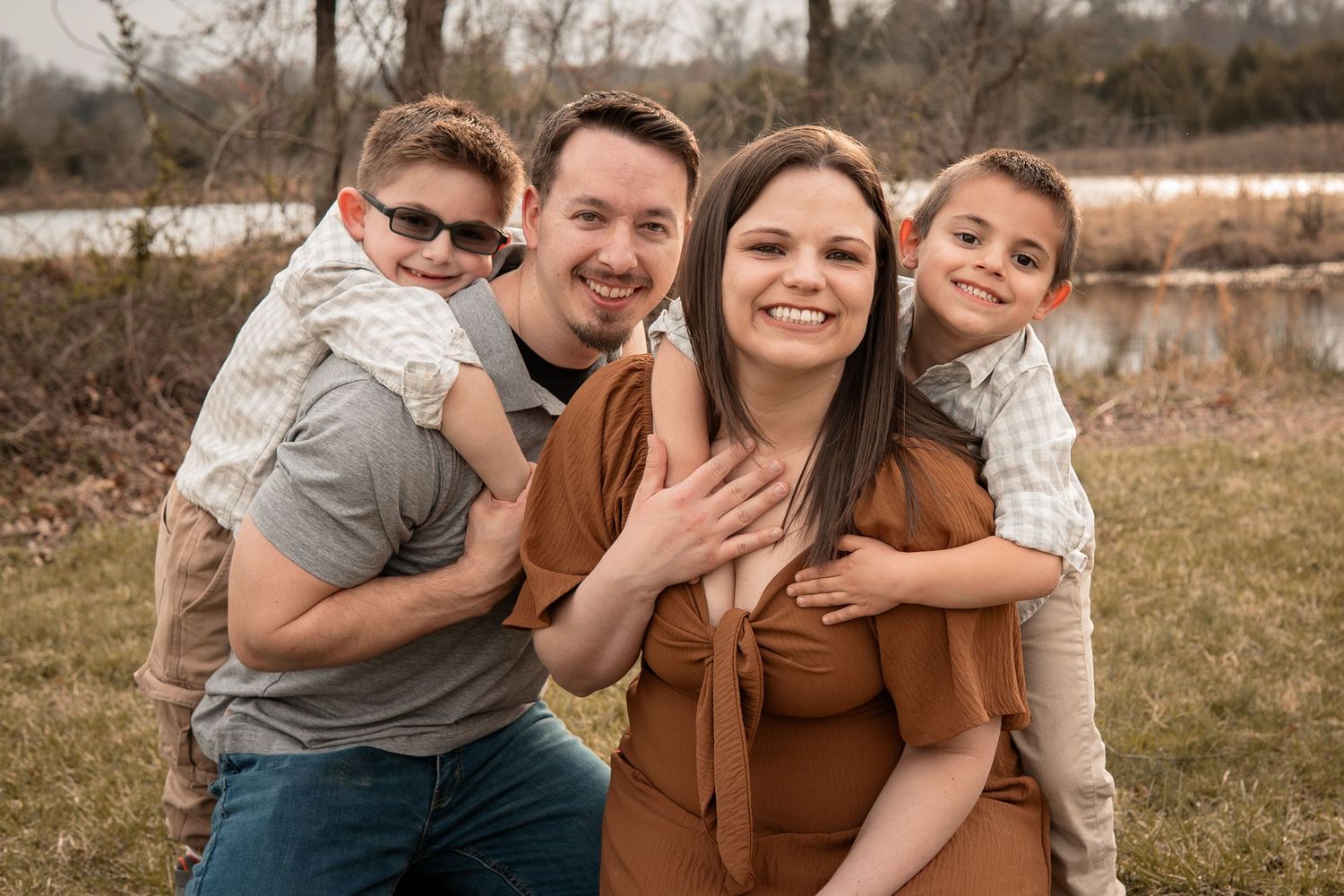 Happy family of four posing outdoors near a pond, parents holding two young boys, smiling in casual autumn attire.