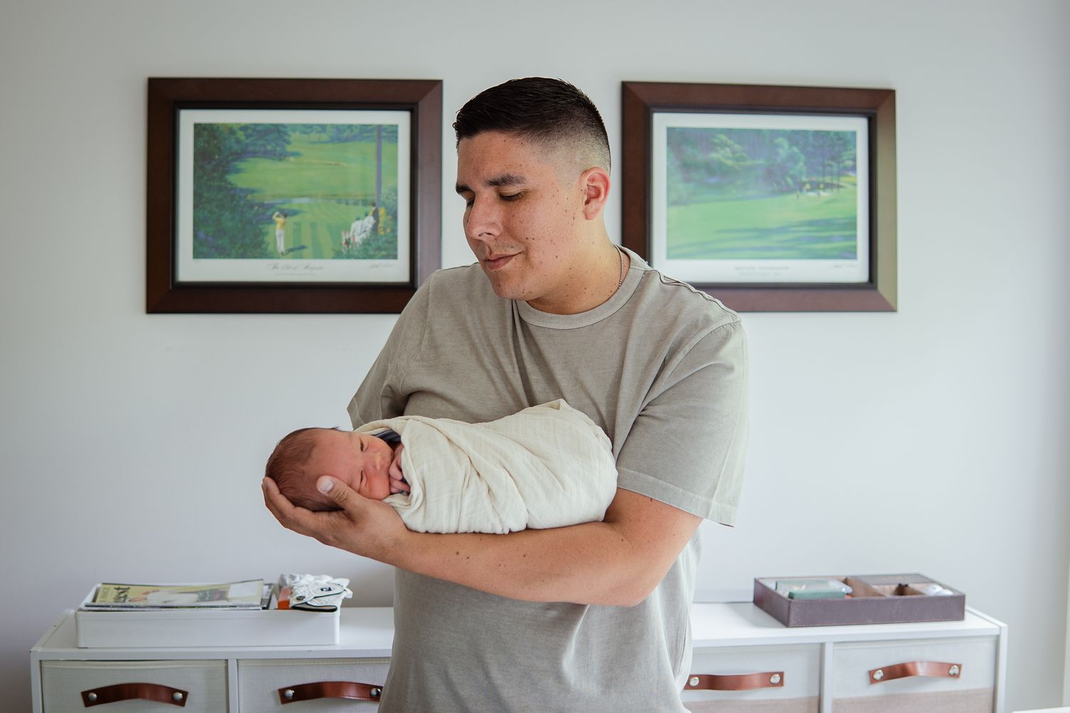 Parent in gray shirt holding swaddled newborn in nursery with framed artwork.