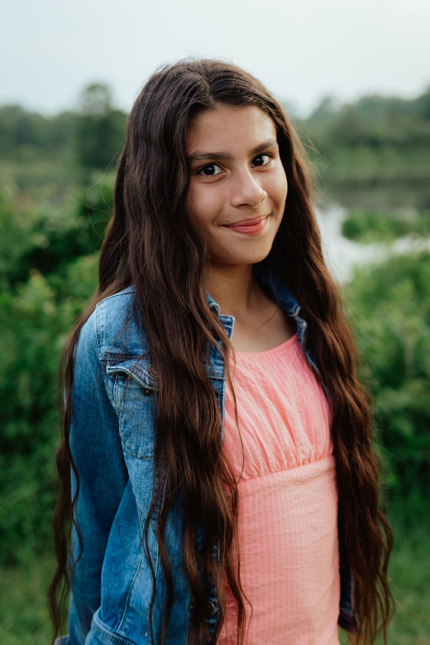 Portrait series of someone with long dark hair wearing a coral top and denim jacket in a natural setting.