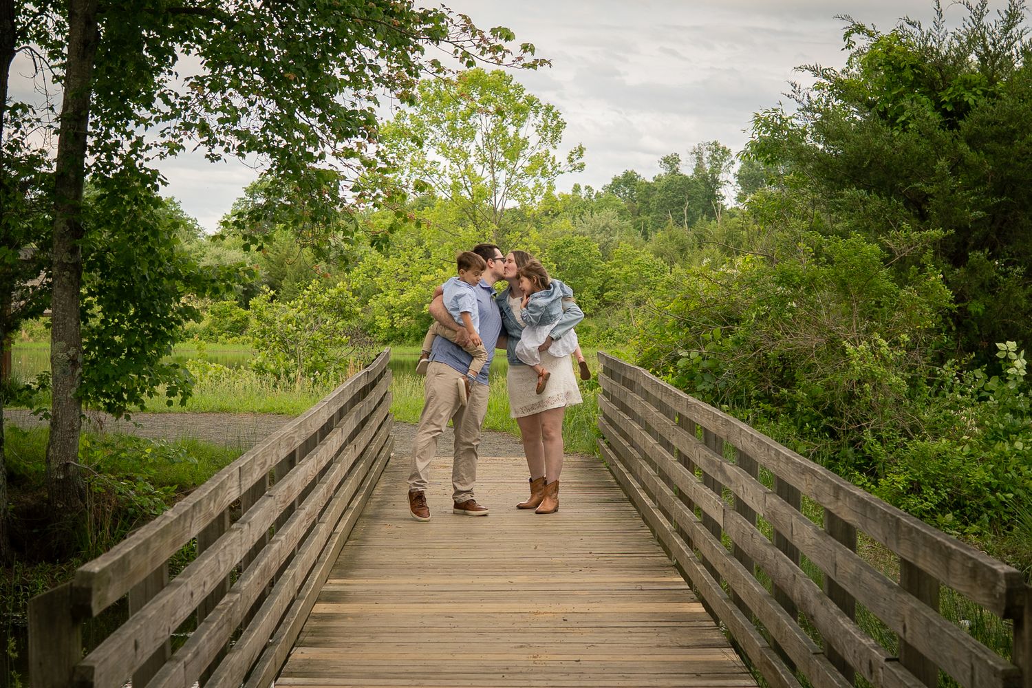 A couple shares a tender moment while standing on a wooden footbridge in a natural parkland setting.