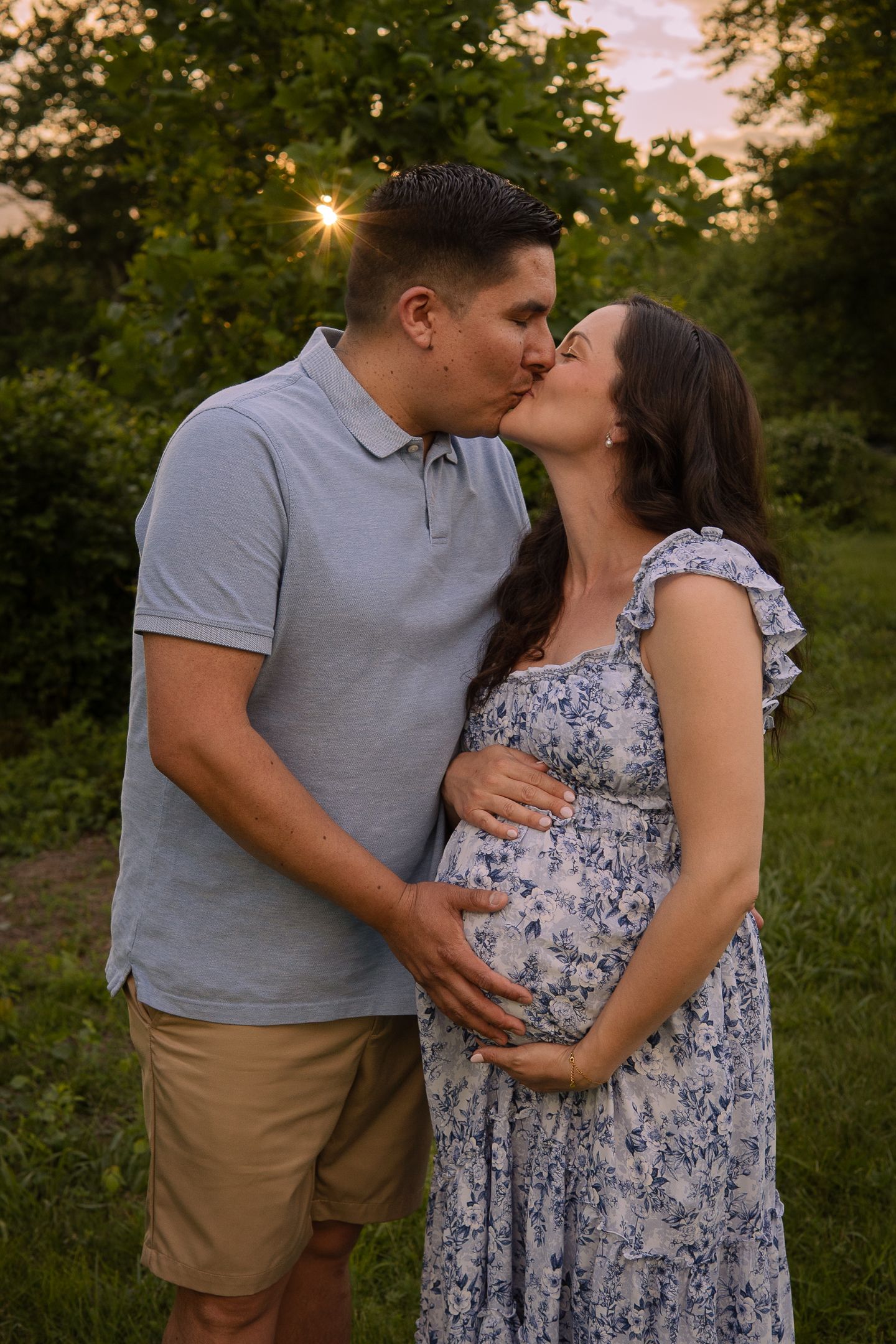 A romantic sunset maternity photo shows a couple in casual attire embracing in a garden setting with sunburst behind trees.