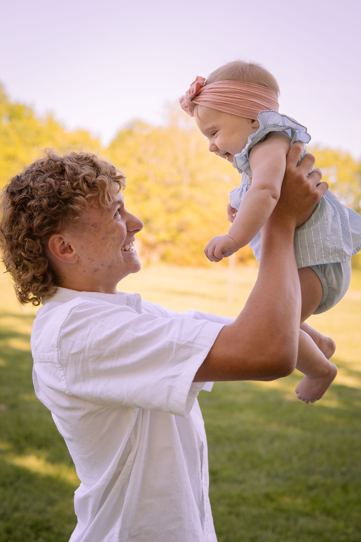 A sweet moment between a young person holding a baby up high against a golden sunset background.
