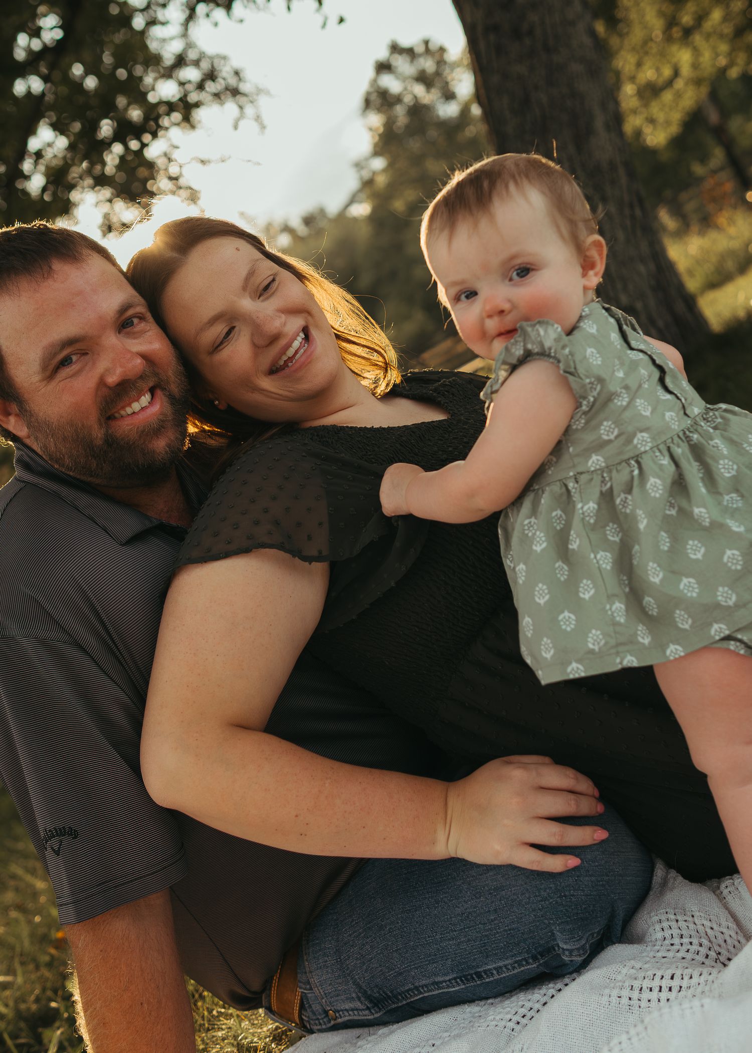 A happy family with a baby in a green dress sitting together on a blanket outdoors at sunset.