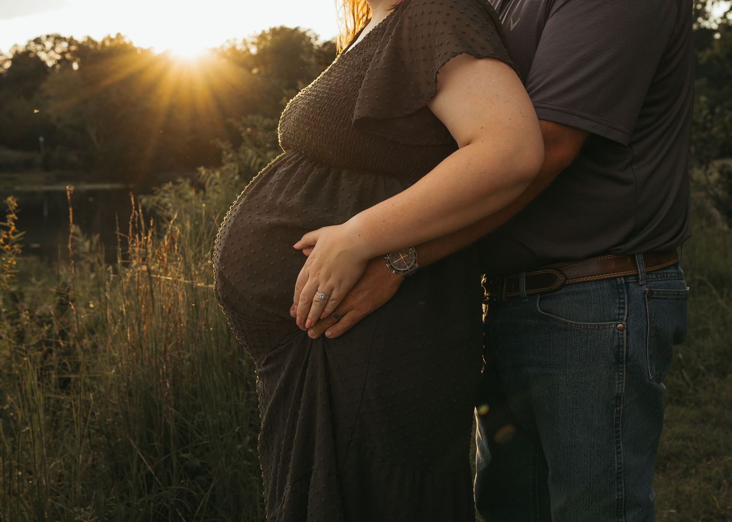 Couple embracing pregnant belly at golden sunset in field during outdoor maternity photo session.