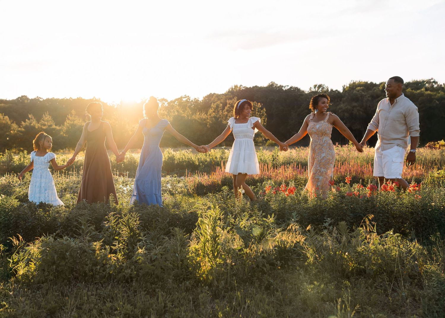 Family holding hands in sunlit meadow at golden hour, wearing white and pastel dresses.