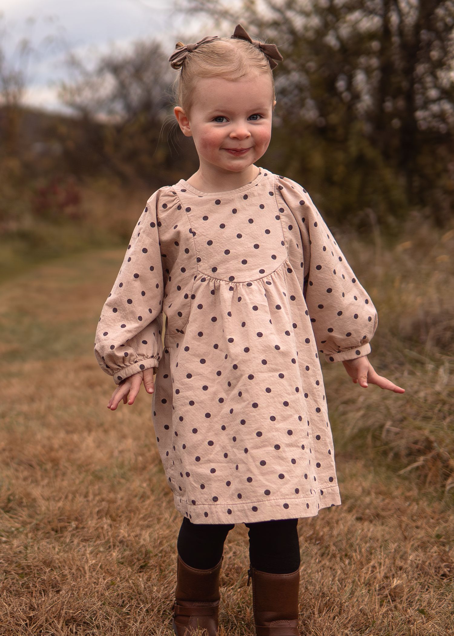 Toddler girl in a pink polka dot dress with brown boots and black leggings, smiling in an autumn field.