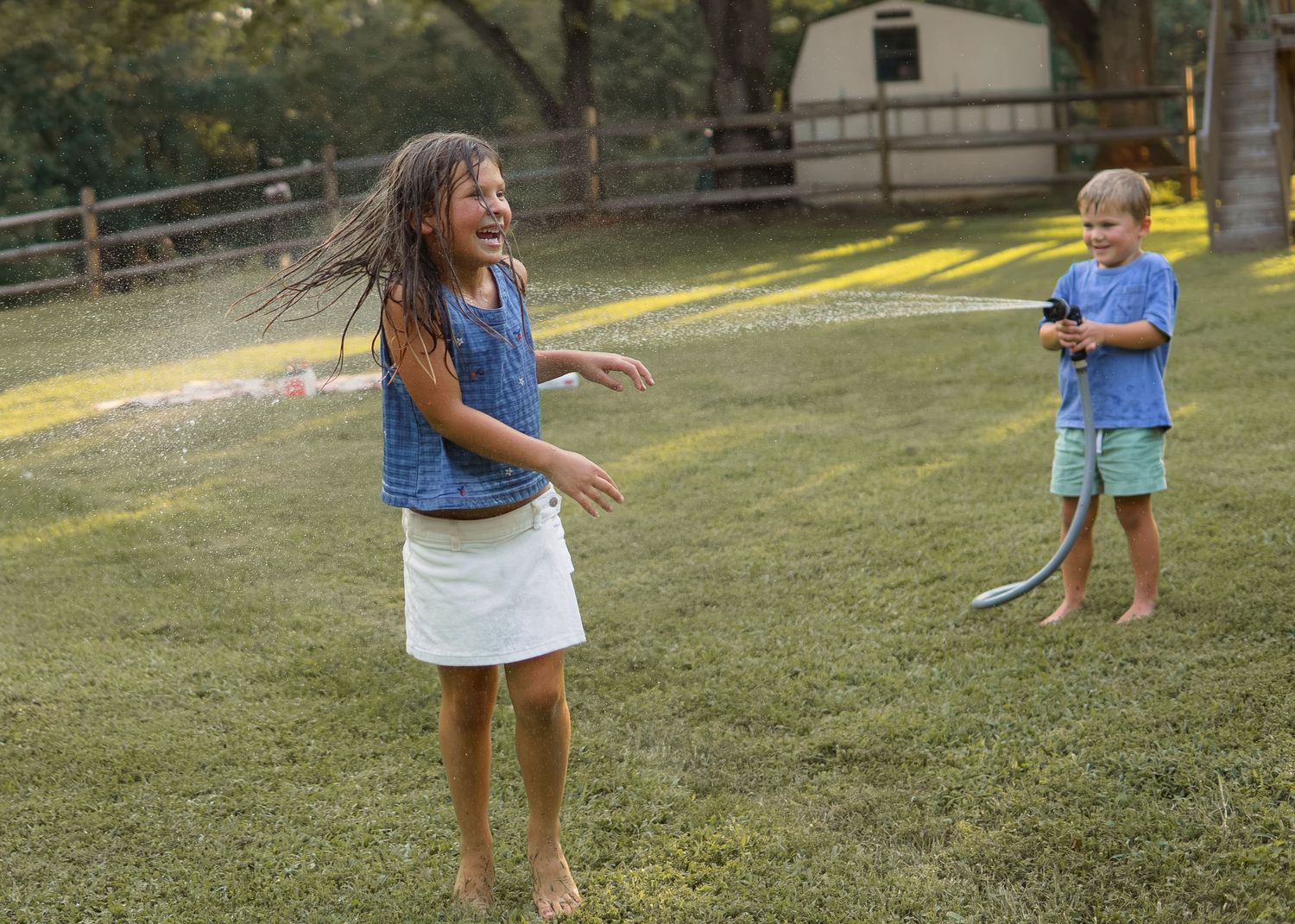 Two children playing in a backyard sprinkler on a summer day, girl laughing in blue top and white skirt.