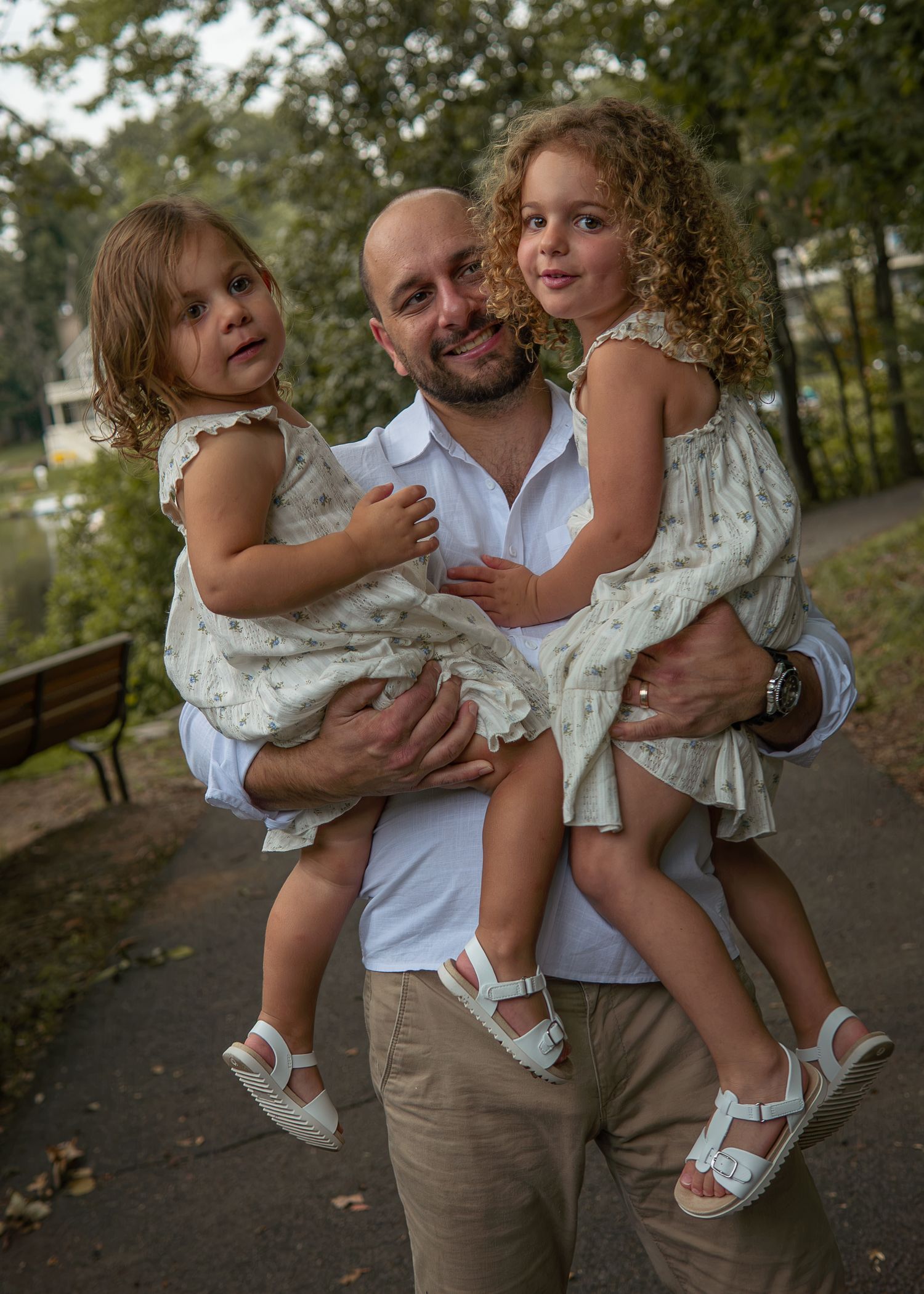Bald man in white shirt holds two young girls in matching floral dresses in a green park setting.