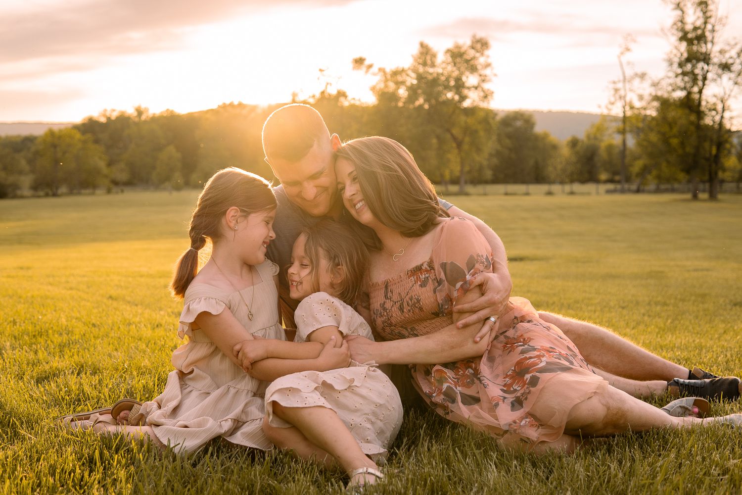 Family in matching pink outfits sitting together on grassy hill during sunset with golden light shining behind them.