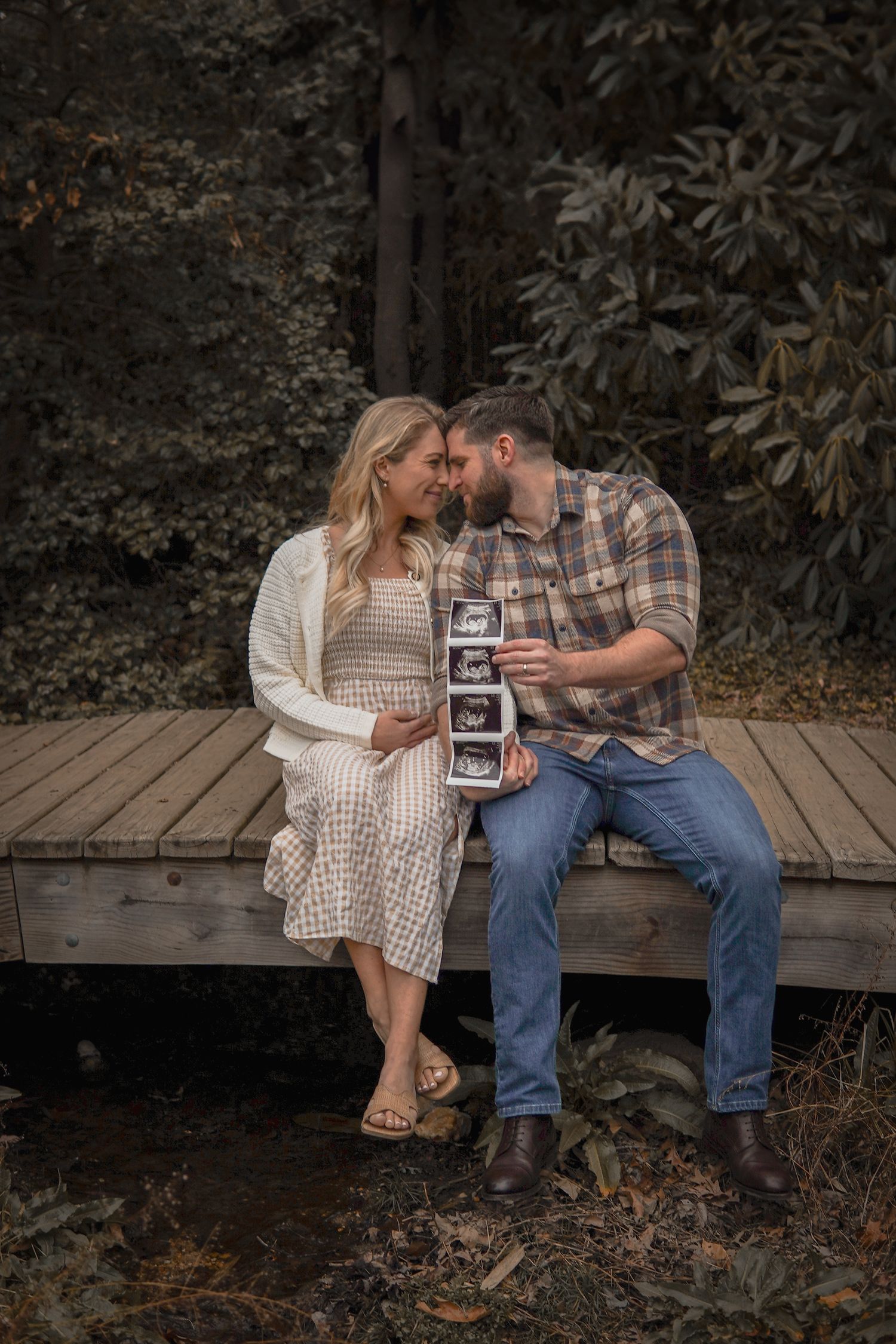 A couple sits intimately on a wooden deck holding ultrasound photos while sharing a tender moment in a moody outdoor setting.