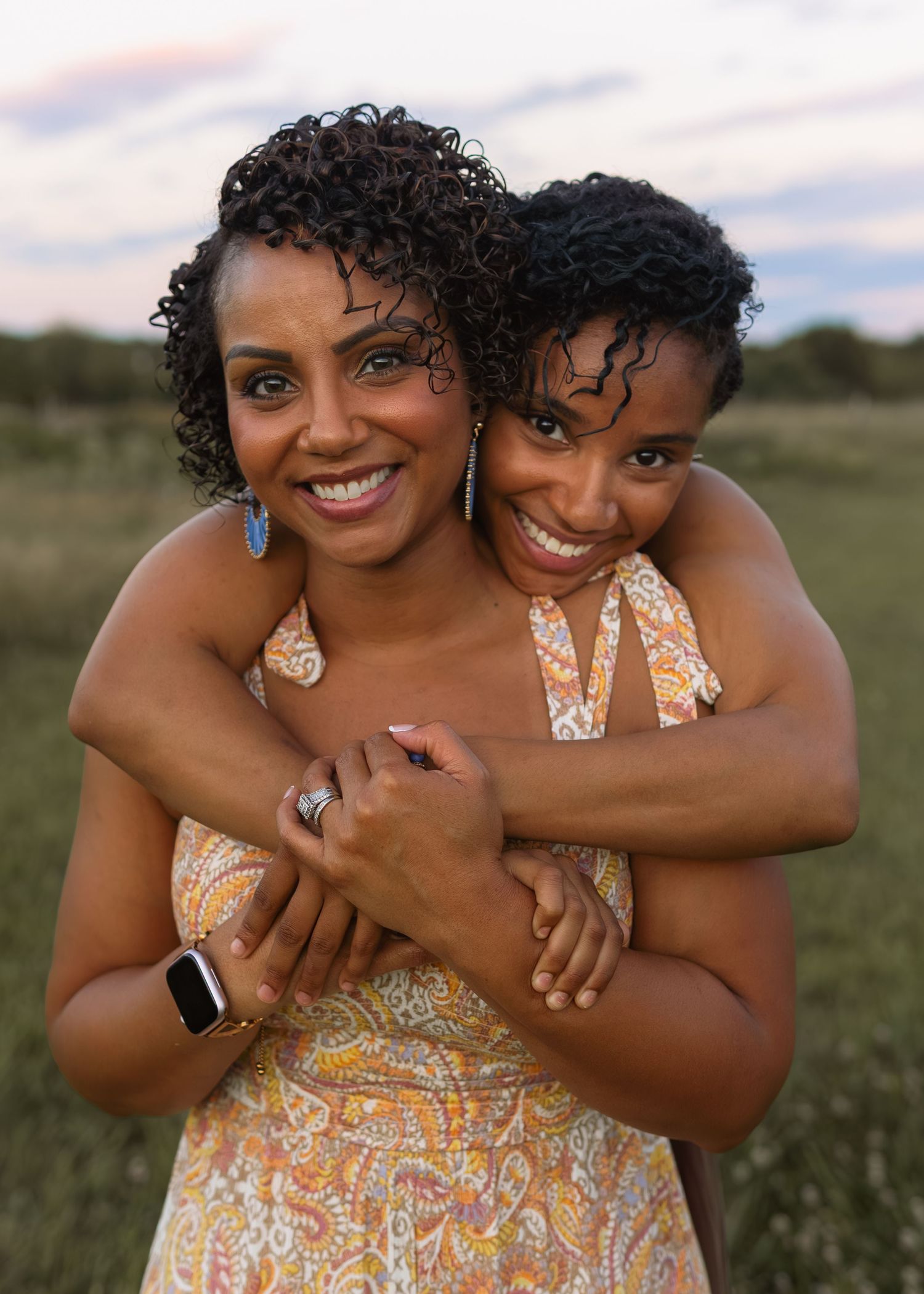 Two Black women smile warmly while embracing in a field during golden hour, wearing summer dresses.