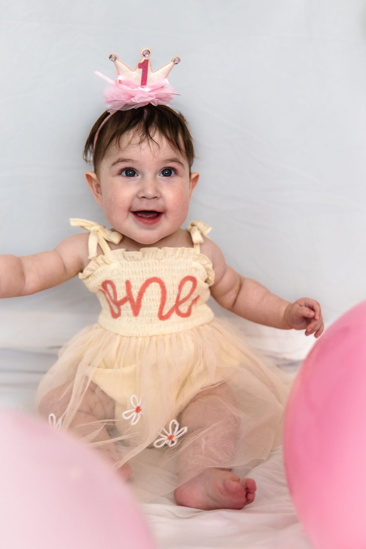 A first birthday celebration with a pink crown and cream tulle dress featuring 'one' text.