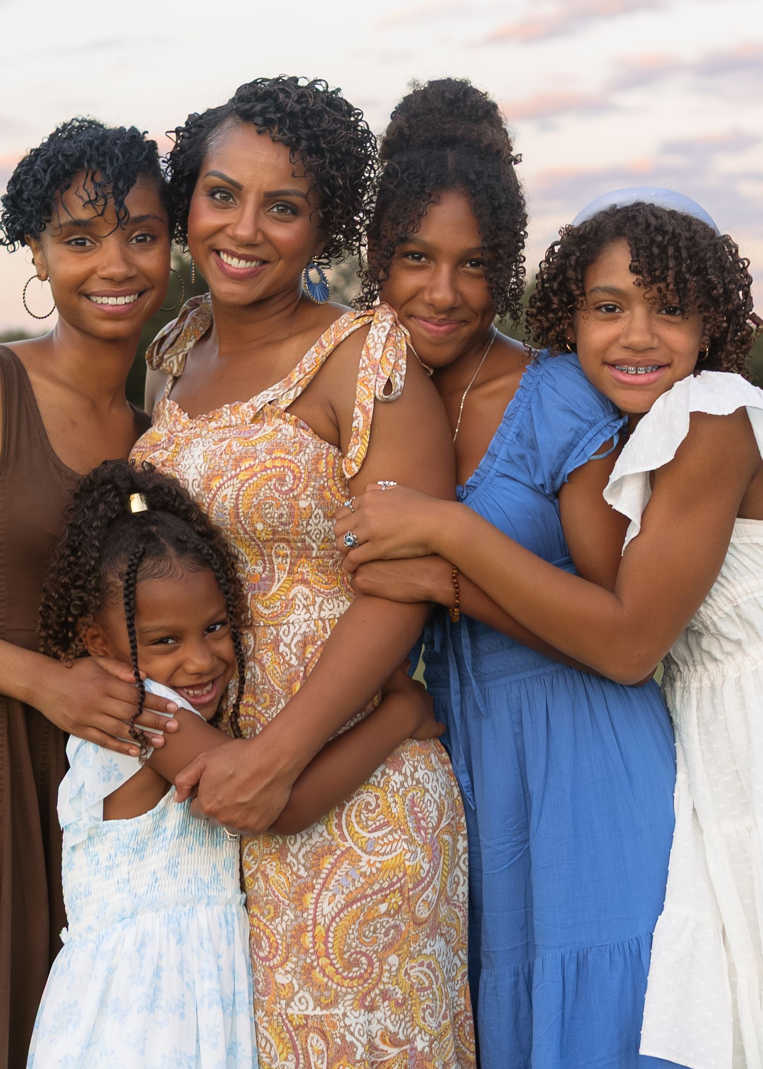 A joyful Black mother poses with her four daughters in a warm outdoor family portrait at sunset.