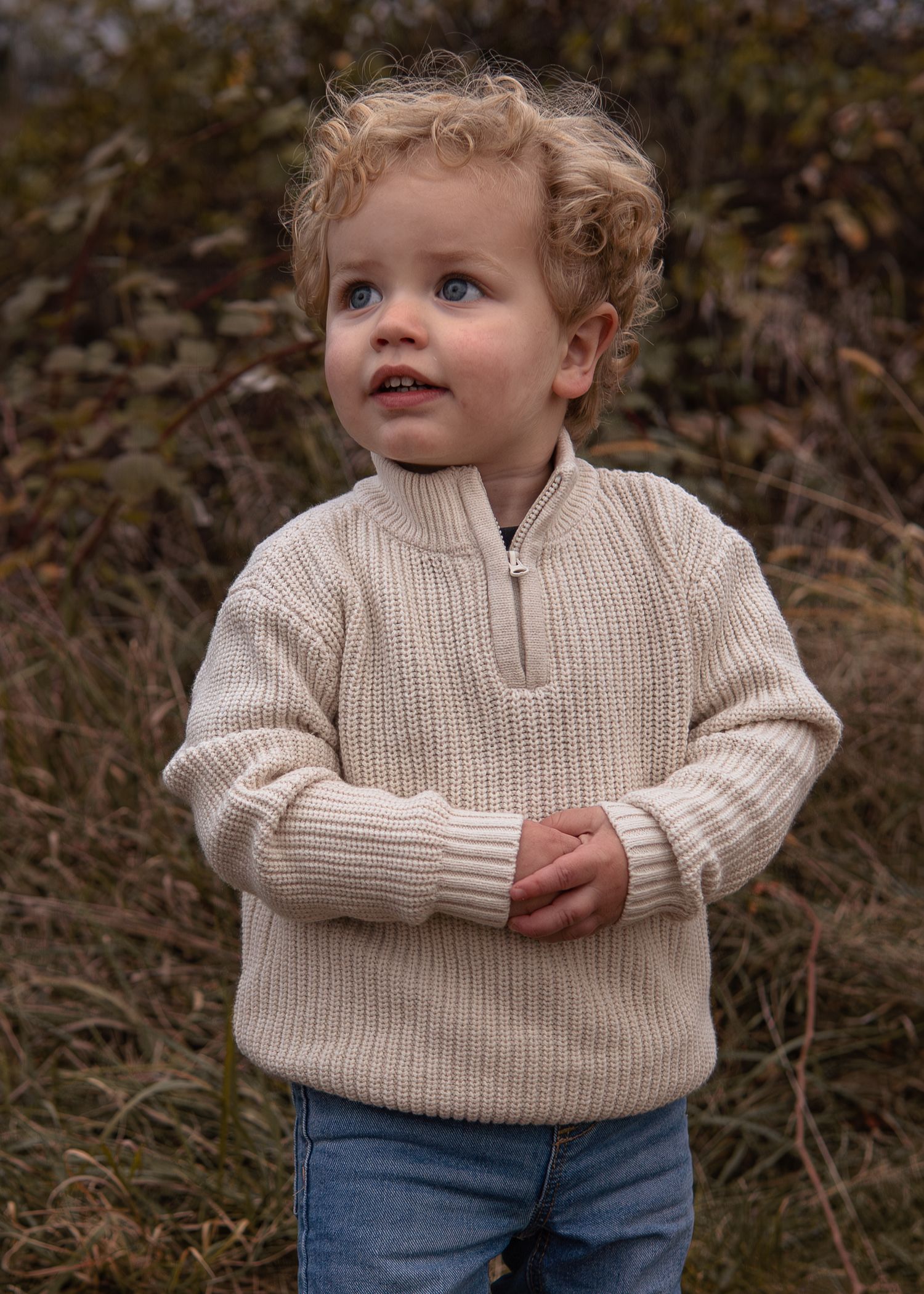 Curly-haired toddler boy in a beige zip-up knit sweater and jeans standing outdoors among green foliage.