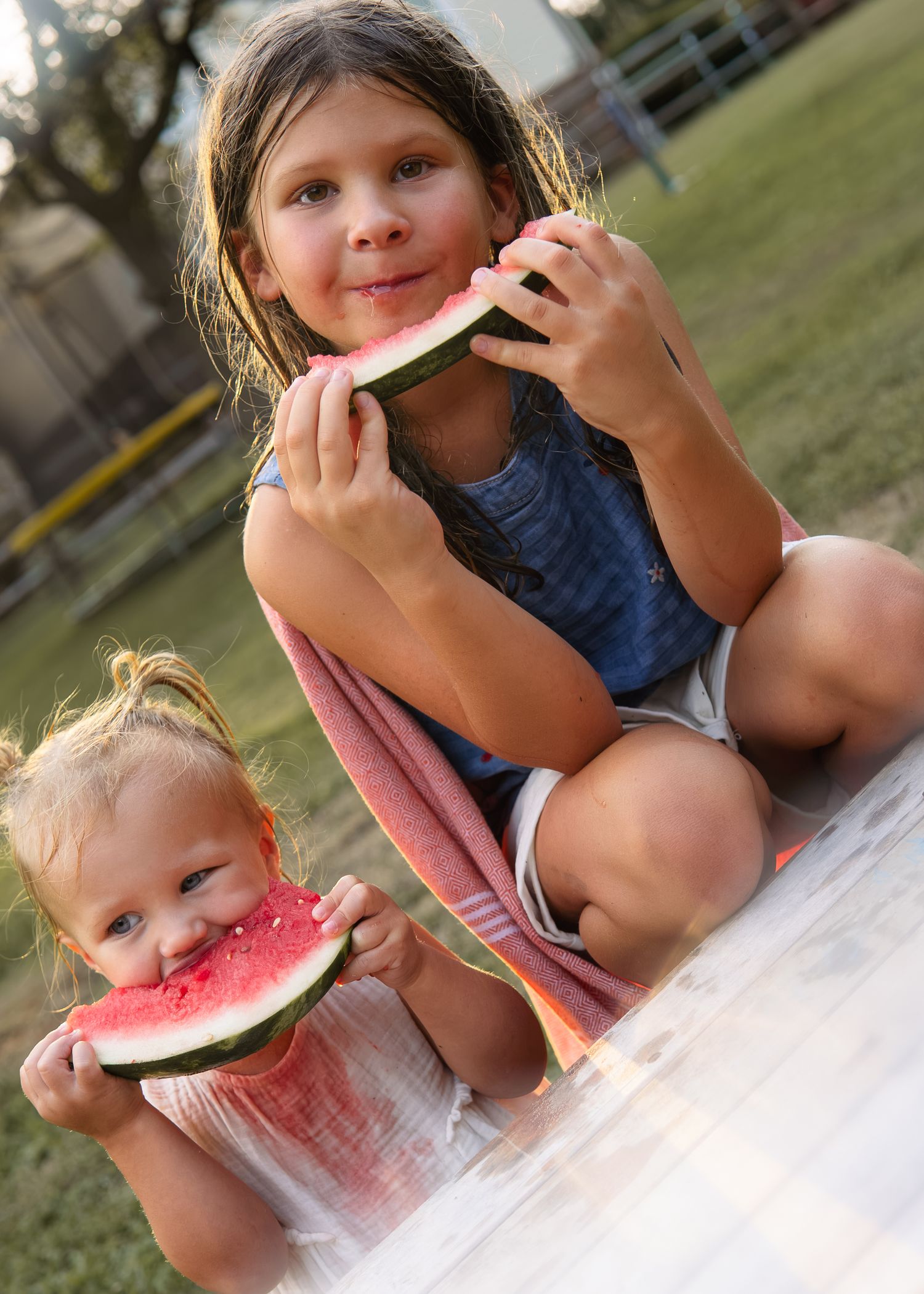 Two young sisters enjoying fresh watermelon slices outdoors, older girl in denim overalls crouching behind toddler.