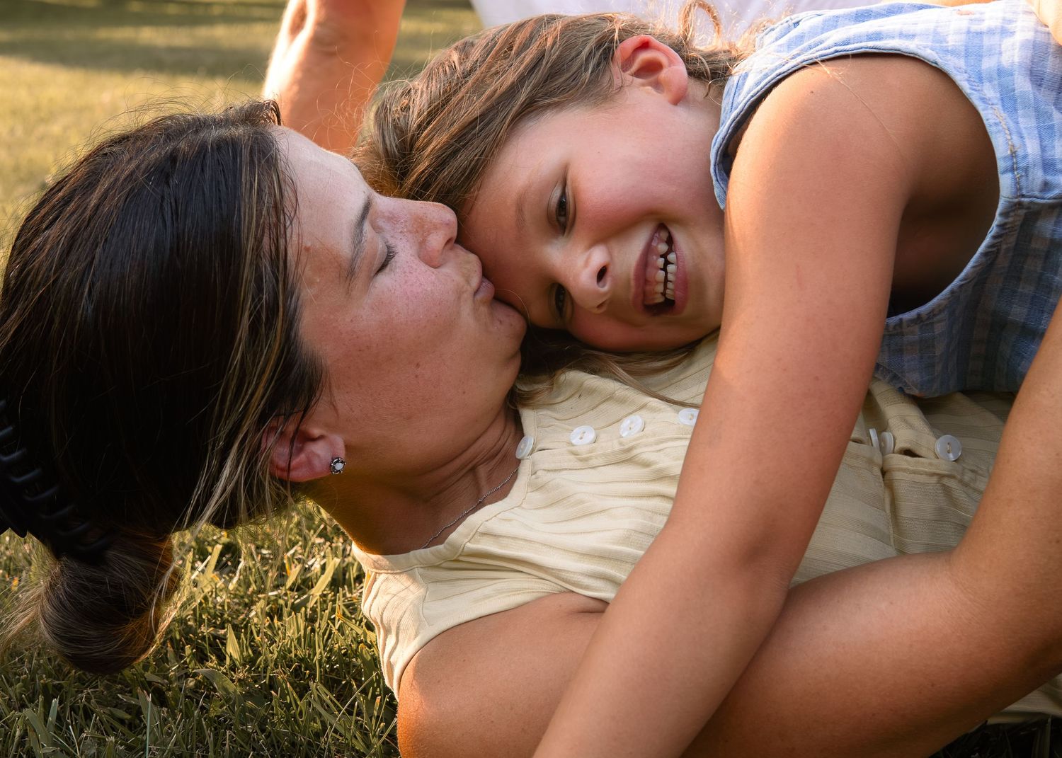 A mother and young daughter share a joyful moment laughing together while lying on green grass outdoors.
