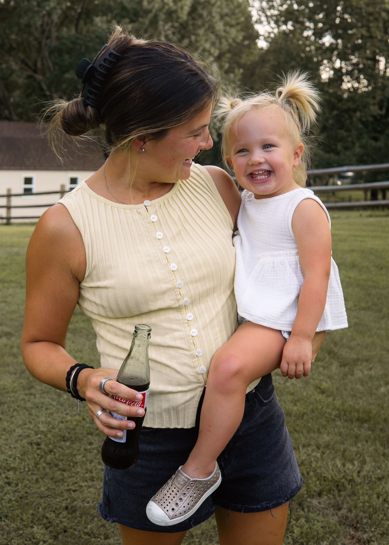 A young woman holds a smiling toddler girl while holding a soda bottle outdoors on a grassy lawn.