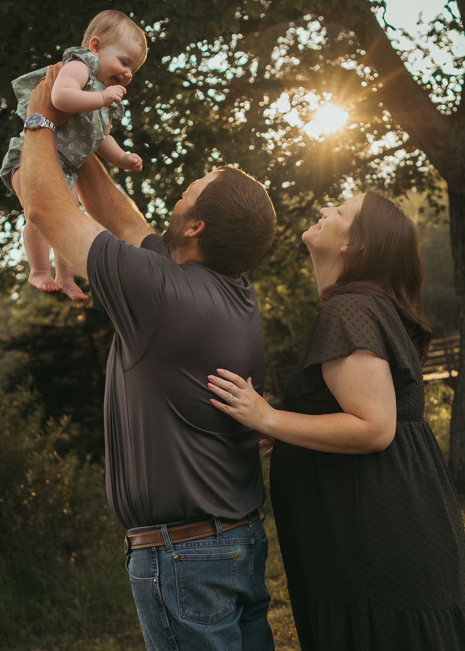 Father lifts smiling baby in golden sunset light while mother watches lovingly outdoors.
