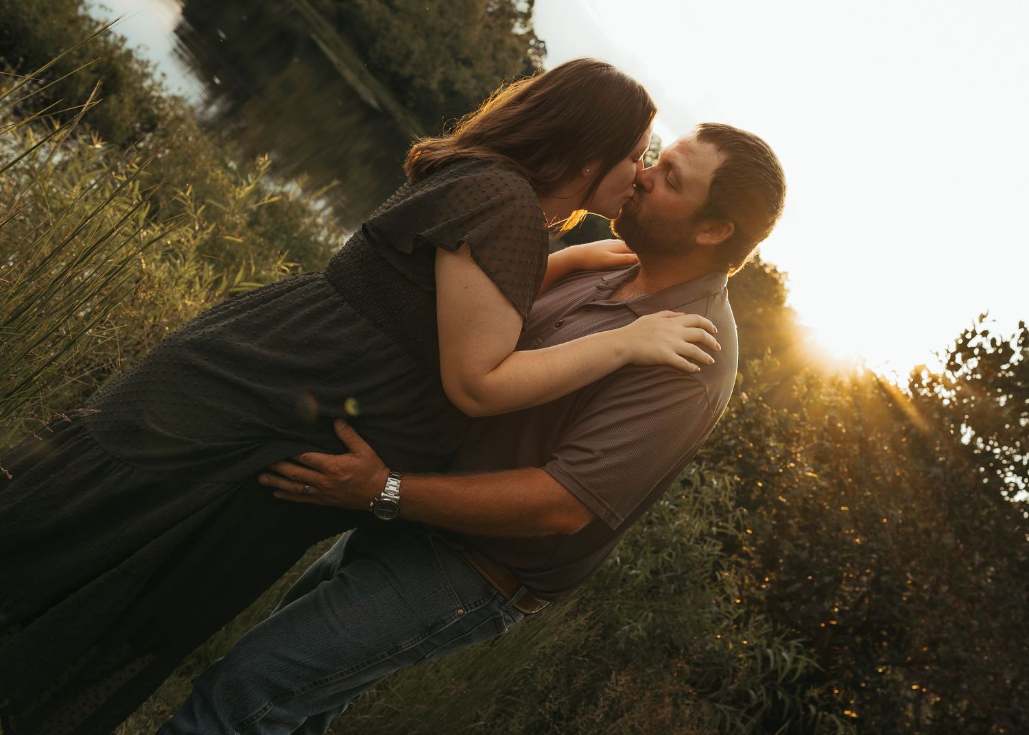 Couple sharing a romantic kiss outdoors at golden hour, embracing near lush greenery with warm sunlight glowing behind them.