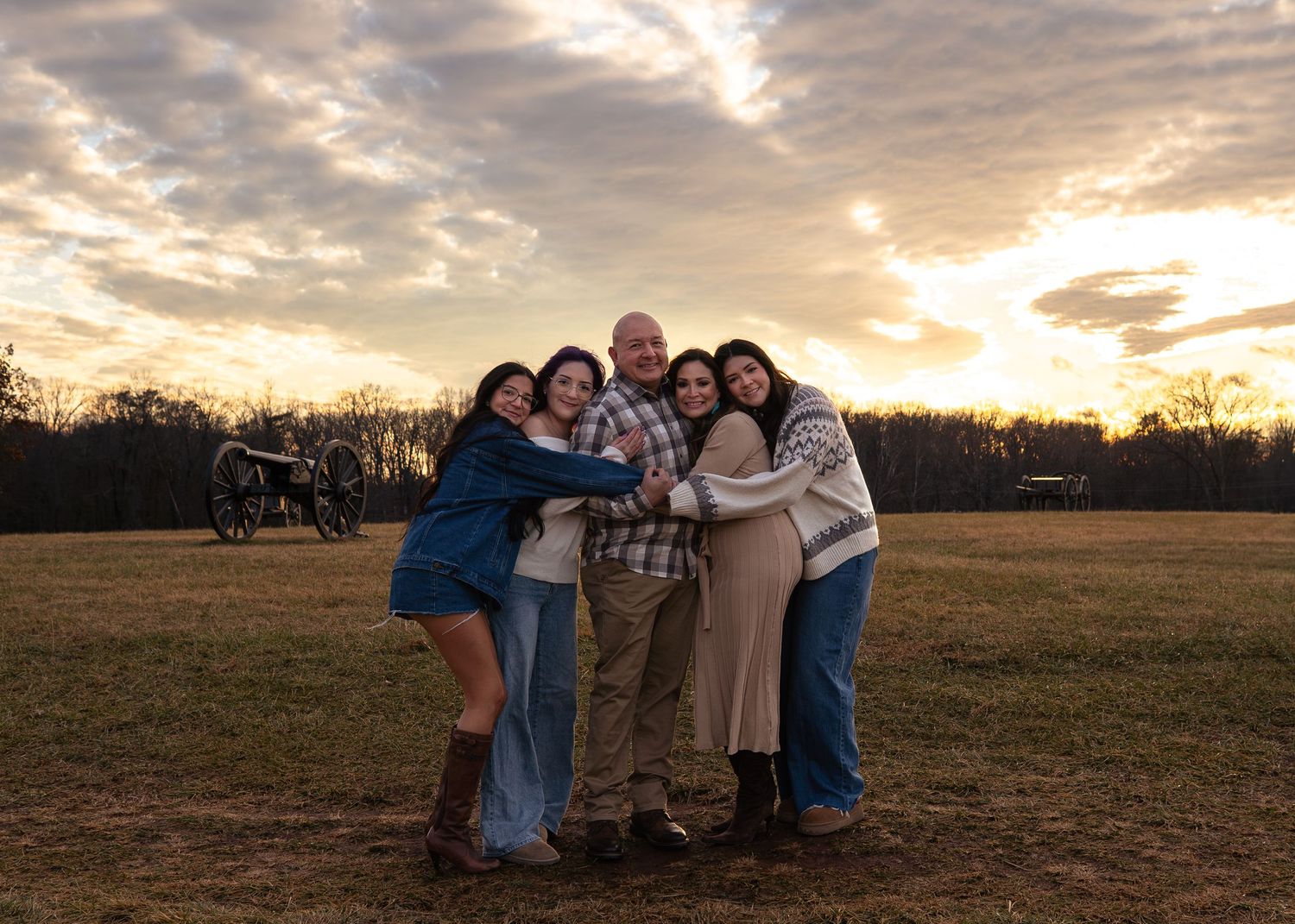 Family of five smiling and hugging at sunset on a historic battlefield with cannons in the background.