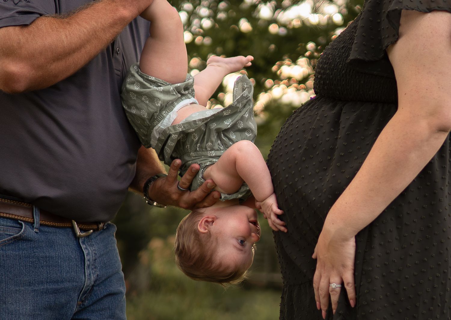 Man holding toddler upside down while pregnant woman stands nearby outdoors during golden hour.